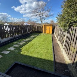 HEKTAR - Zadbany ogród z równo przystrzyżoną trawą, ścieżką z płyt, drewnianym domkiem narzędziowym i rabatami wzdłuż ogrodzenia. Błękitne niebo z chmurami w tle.