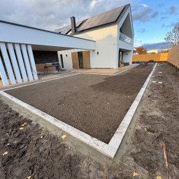 HEKTAR - Przygotowany teren pod trawnik z obrzeżem z kostki przy nowoczesnym domu z panelami słonecznymi i drewnianym ogrodzeniem. Widoczne narzędzia i deski.