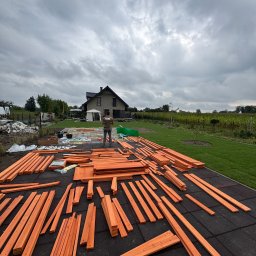 HEKTAR - Przygotowane elementy drewniane na czarnej, gumowej nawierzchni, prawdopodobnie do budowy konstrukcji ogrodowej. W tle dom i zielony trawnik pod pochmurnym niebem.