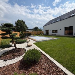 HEKTAR OGRODY & BRUK - Nowoczesny ogród z basenem, zadbanym trawnikiem i strefami z białym żwirem oraz korą. W tle biały dom, pergola i formowane rośliny. Słoneczne ujęcie.