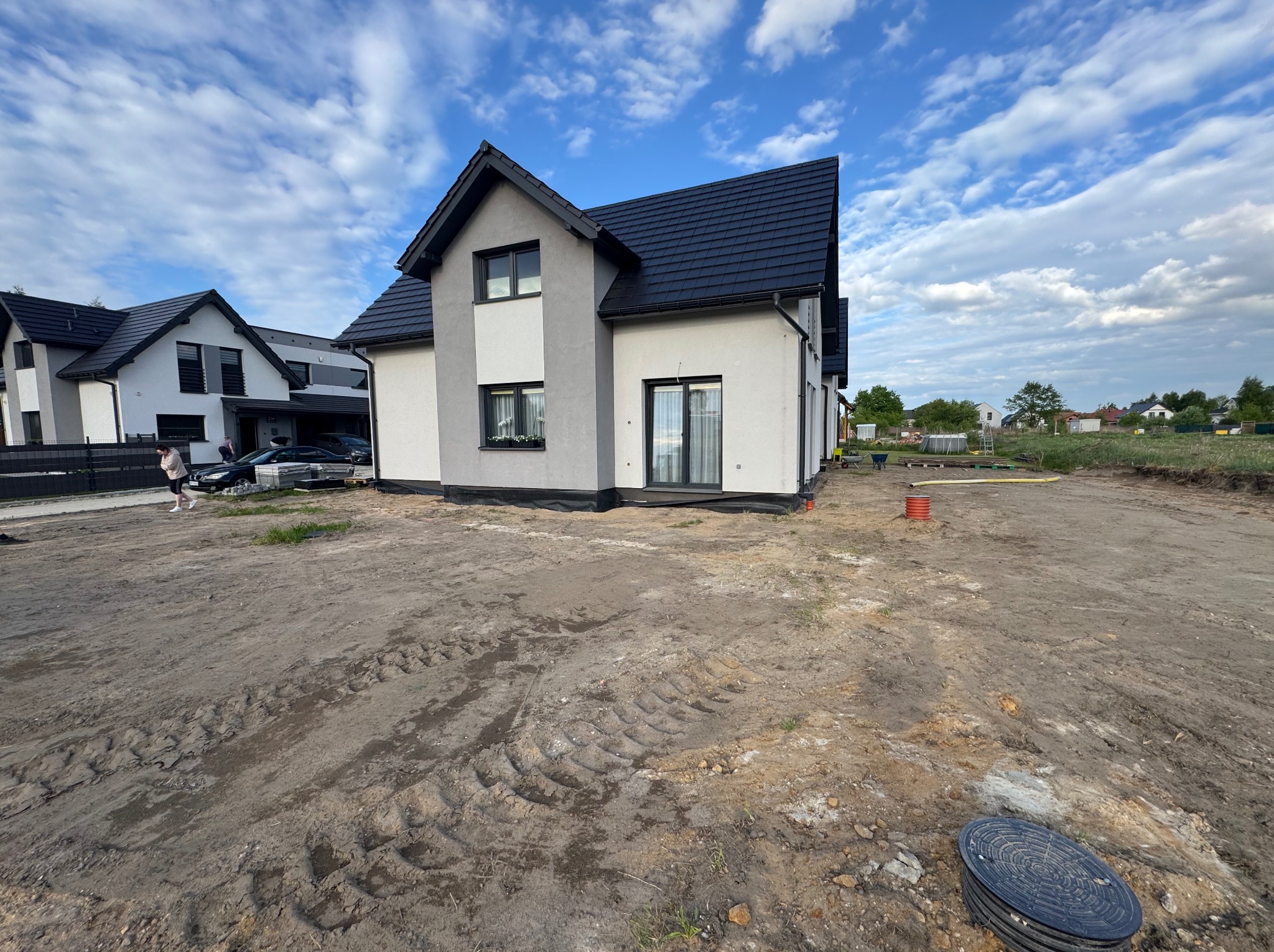 Nowoczesny dom jednorodzinny z czarnym dachem, widok z terenu przygotowanego pod ogród. Ślady opon na ziemi. Błękitne niebo z chmurami w tle.