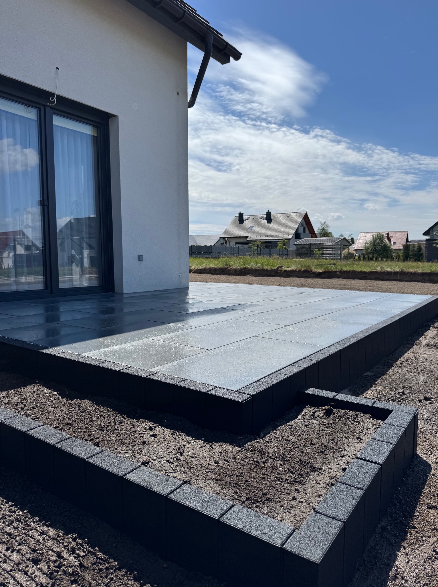 Nowoczesny taras z szarych płyt, obrzeża z czarnej kostki. Widok na dom i błękitne niebo z chmurami.