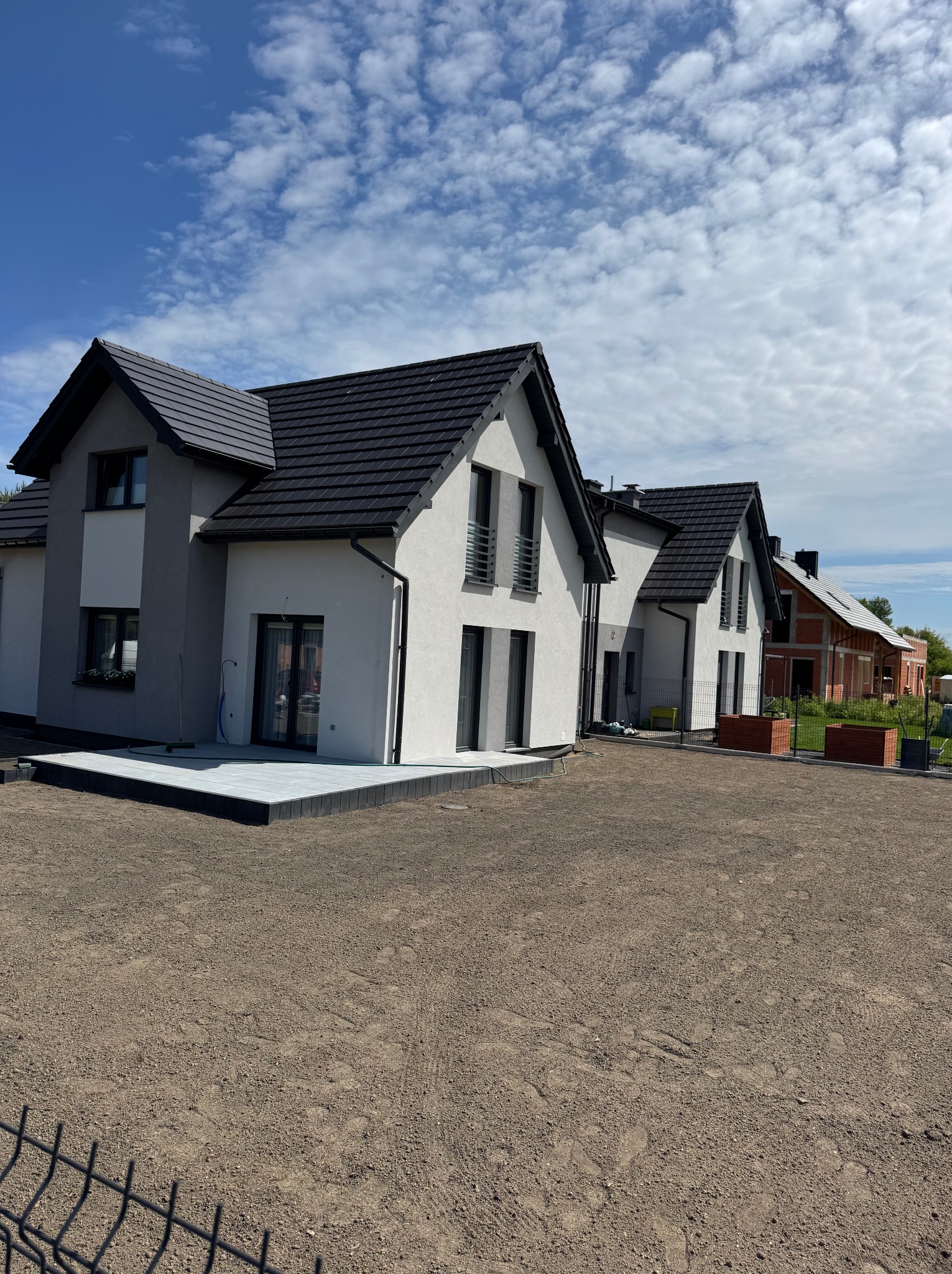 Nowoczesne domy jednorodzinne z szarą kostką brukową na tarasie i przygotowanym terenem pod ogród, widok w słoneczny dzień.