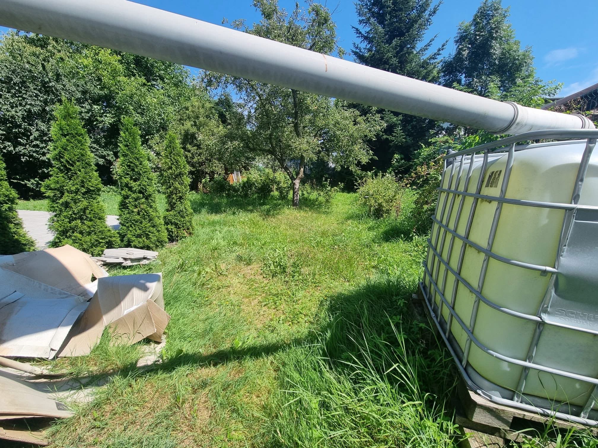 Zielony ogród z systemem nawadniania: duży zbiornik na wodę, rura i bujna trawa. Widoczne drzewa iglaste i liściaste. Częściowo widoczne kartony.