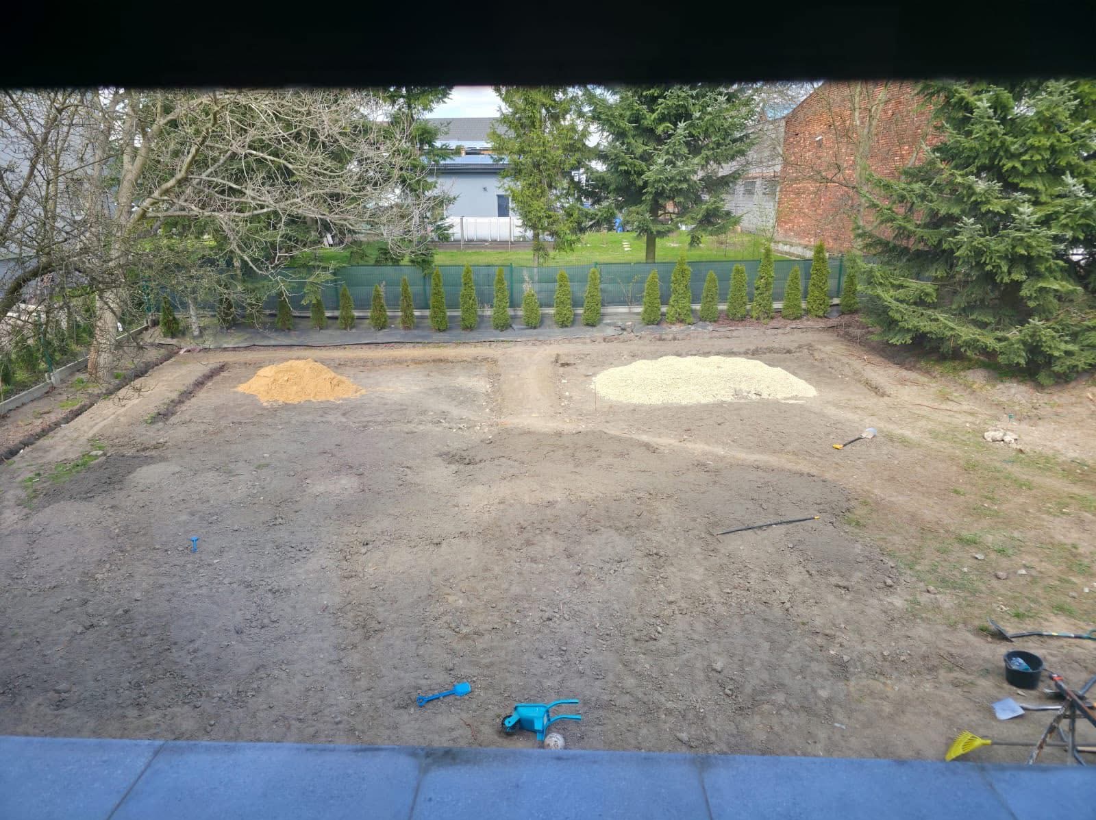 Przygotowany teren pod budowę: wykopane fundamenty, usypane dwie pryzmy piasku i żwiru, widoczne narzędzia ogrodowe. Tło: rząd tuj i zabudowa.