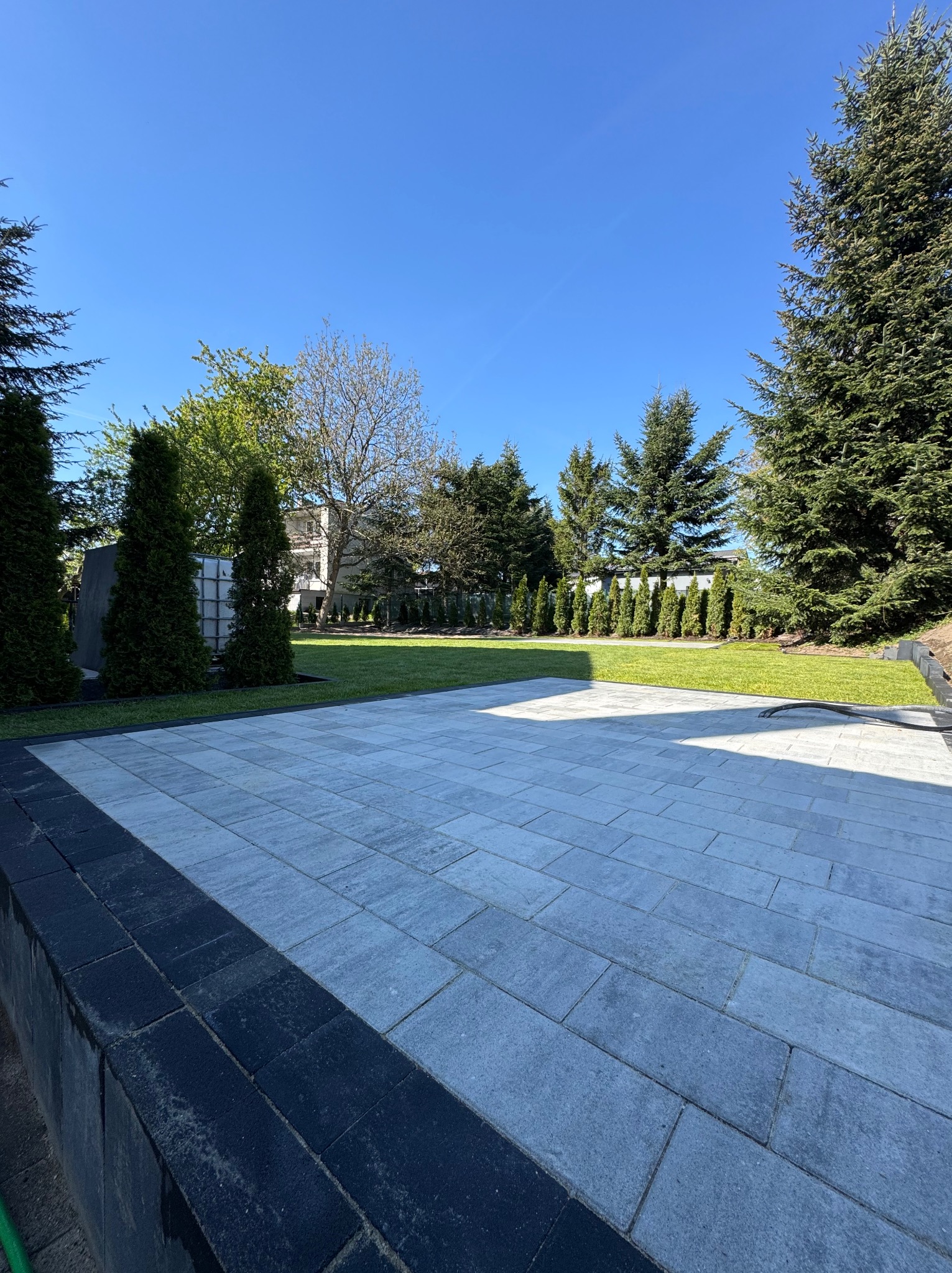 Wyłożony kostką brukową taras z obrzeżem, przylegający do zadbanego trawnika i żywopłotu z iglaków w słoneczny dzień. Widok na ogród.