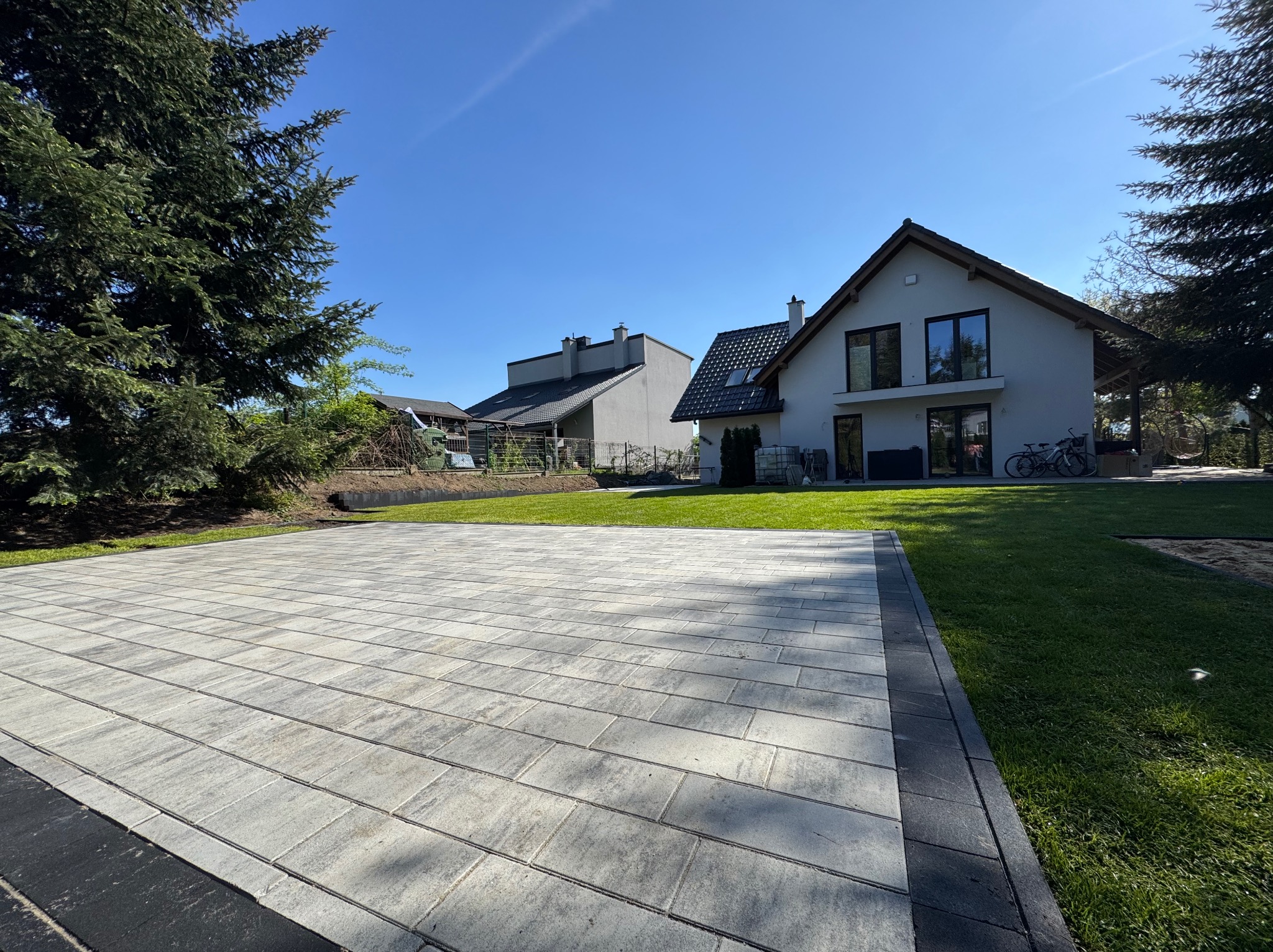 Jasnoszara kostka brukowa ułożona na podjeździe przed domem z zielonym trawnikiem i drzewami w tle, w słoneczny dzień.