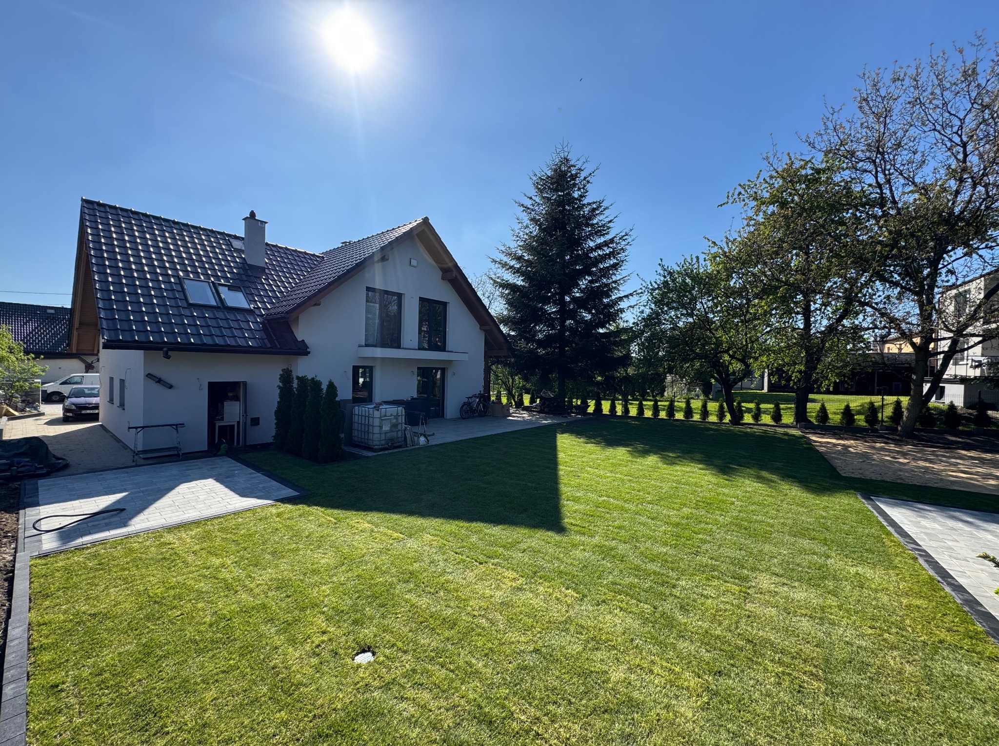 Rozległy, zielony trawnik przed domem z kostką brukową, drzewami i krzewami w słoneczny dzień. Widoczny fragment elewacji budynku.