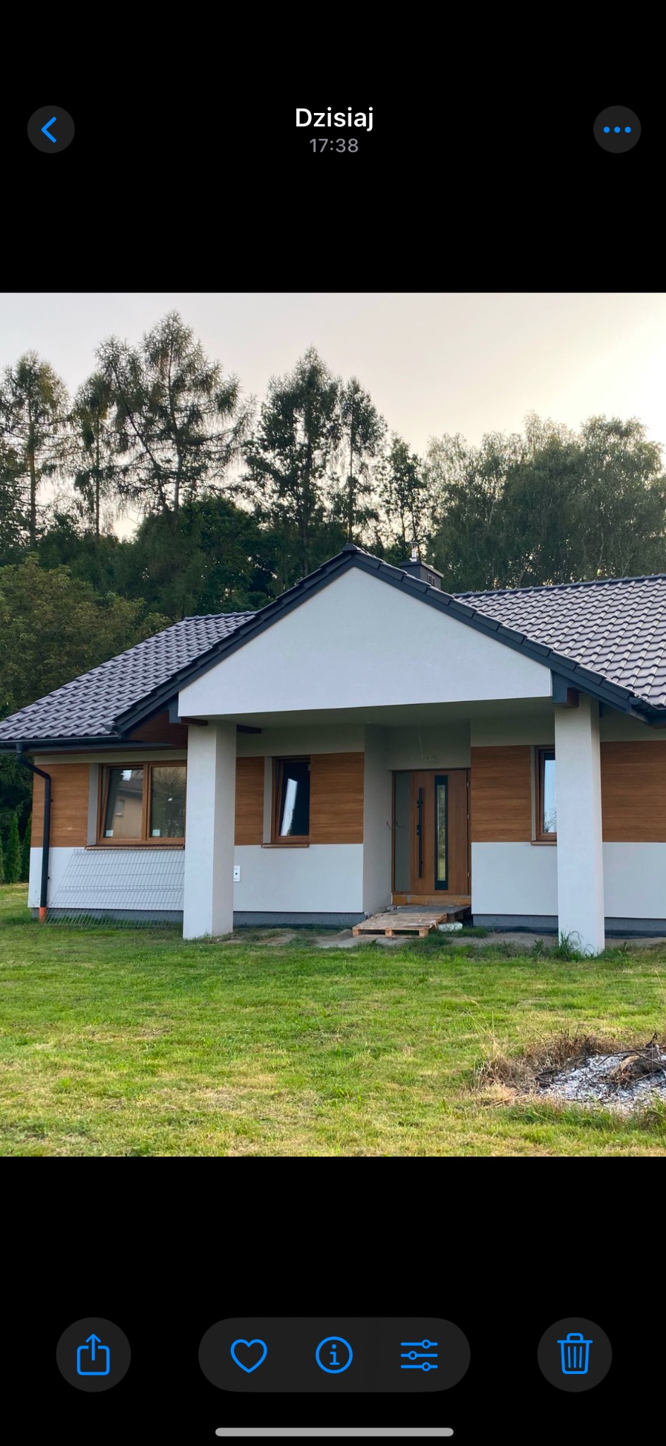 Nowoczesny dom jednorodzinny z drewnianą elewacją i ciemną dachówką, otoczony zielonym trawnikiem, w tle widoczne drzewa. Widoczne palety przed wejściem.