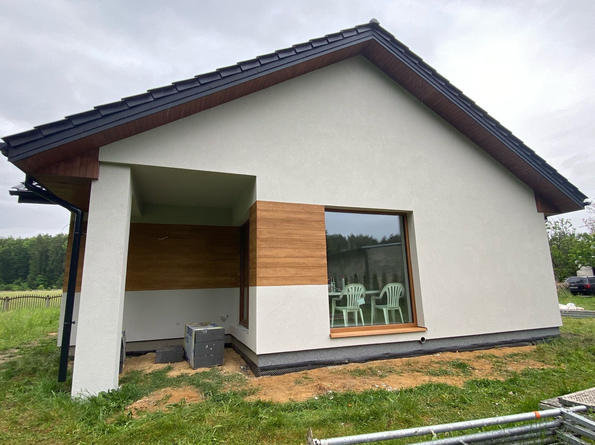 Nowoczesny dom jednorodzinny z drewnianą elewacją i dużym oknem, widok z zewnątrz, w tle zieleń. Elementy wykończeniowe w trakcie realizacji.