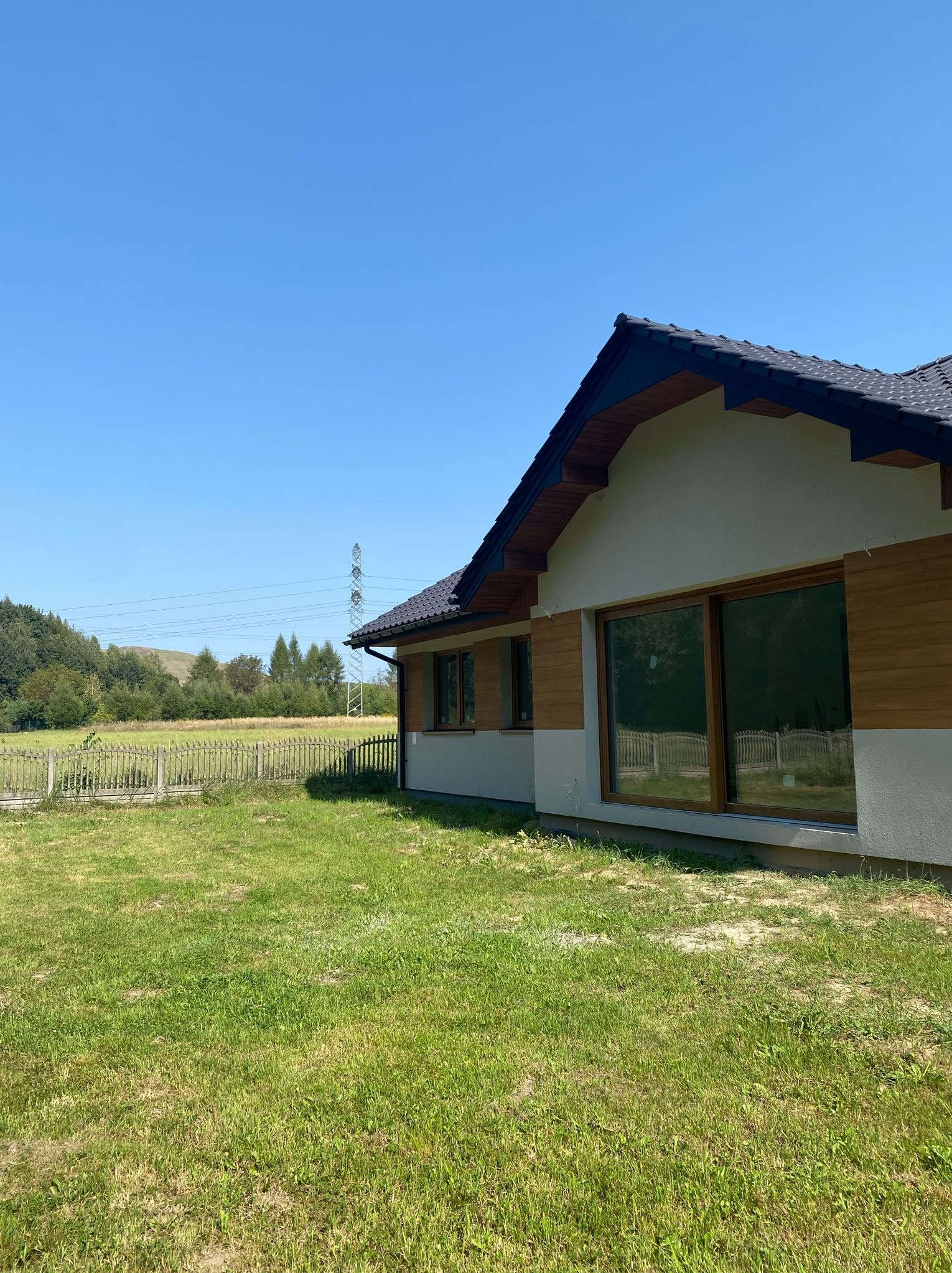 Nowoczesny dom jednorodzinny z drewnianą elewacją i dużymi oknami, otoczony trawnikiem i ogrodzeniem. W tle las i słupy wysokiego napięcia pod błękitnym niebem.