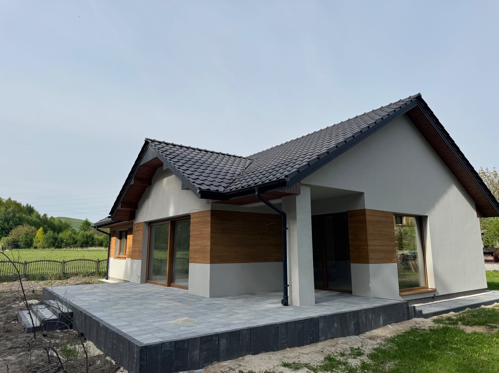 Nowoczesny dom z tarasem wyłożonym szarą kostką brukową i obrzeżem z ciemnych elementów. Widok na elewację z drewnianymi akcentami i zielone otoczenie.