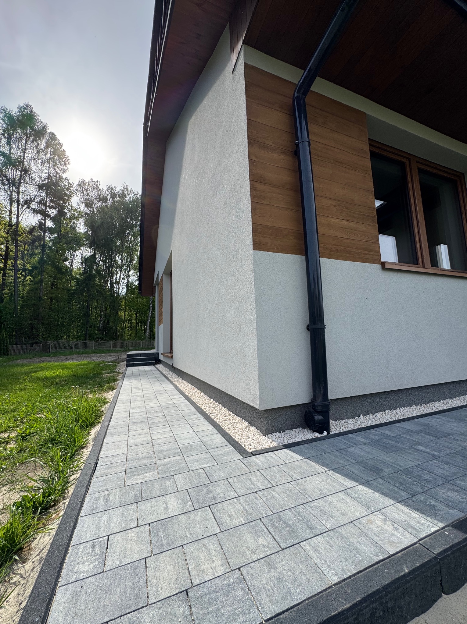 Szara kostka brukowa ułożona przy nowoczesnym domu z drewnianą elewacją, widok z dołu, w tle zieleń. Krawężnik oddziela kostkę od trawnika.