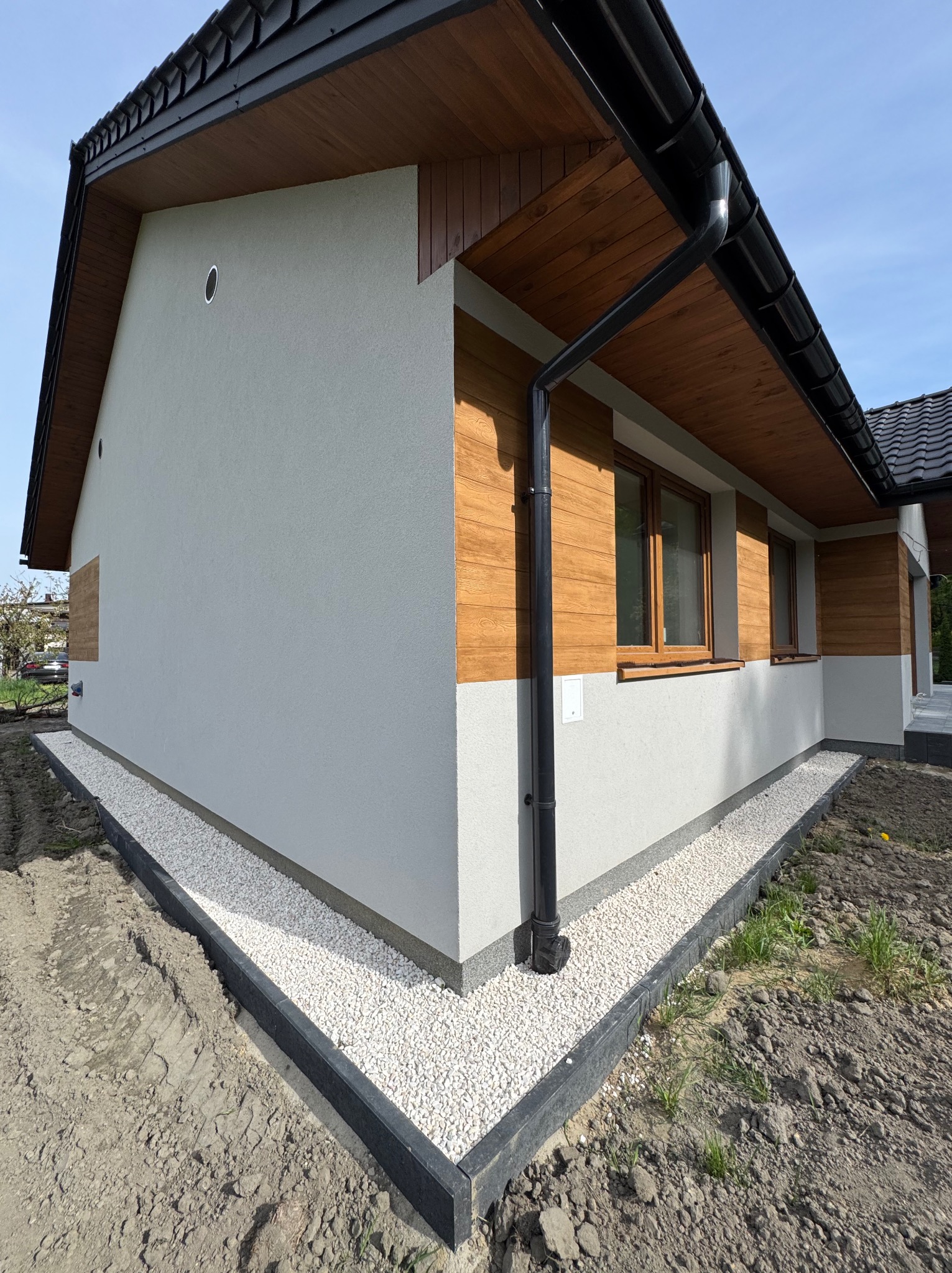 Wykończony fragment elewacji domu z obrzeżem i białym grysem. Widoczne elementy drewniane i czarna rynna. Obrzeże wykonane z ciemnych elementów.