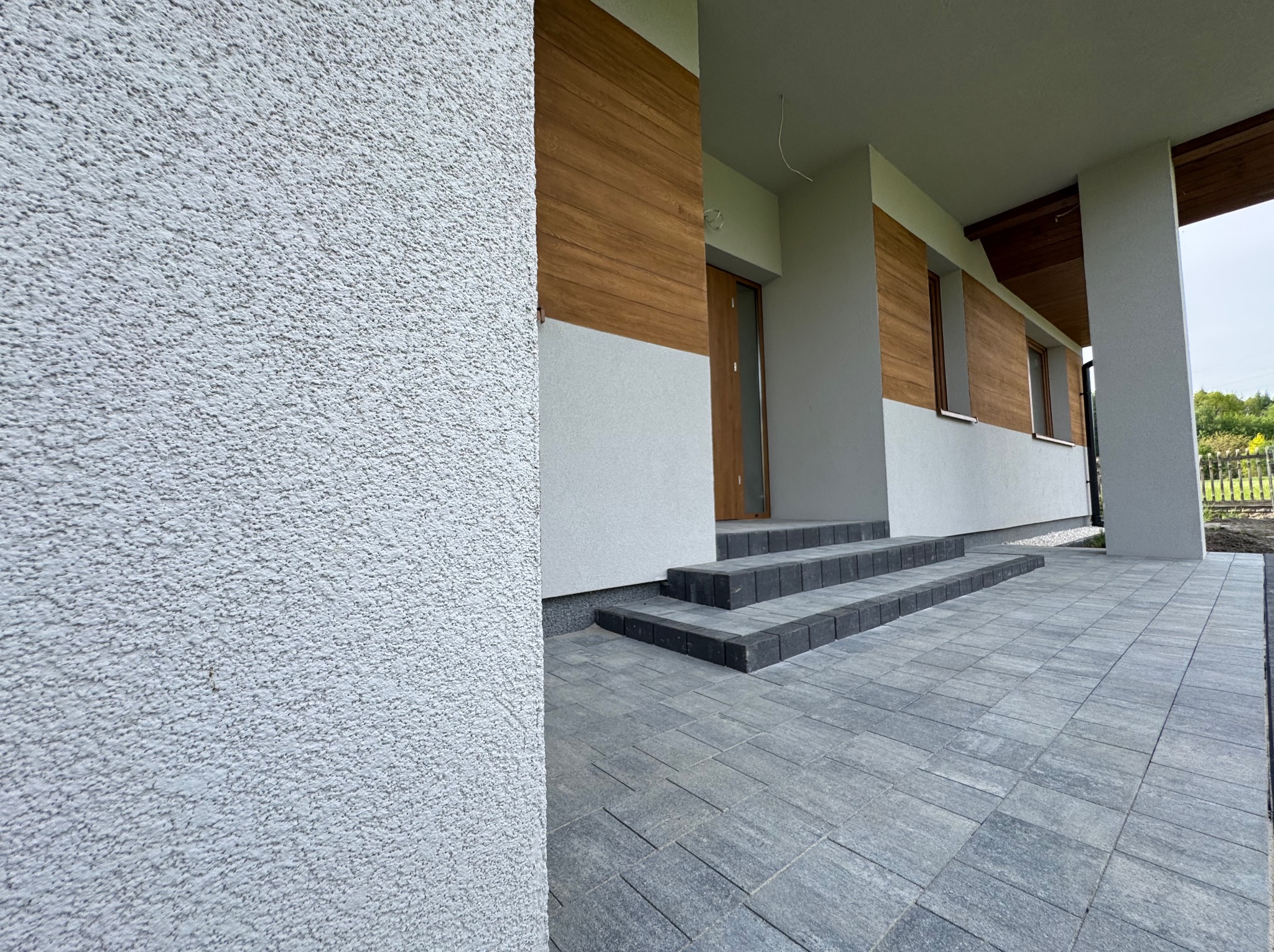 Nowoczesna aranżacja wejścia do domu: szara kostka brukowa, schody z ciemnej kostki, elewacja z elementami drewna. Estetyczne wykończenie.