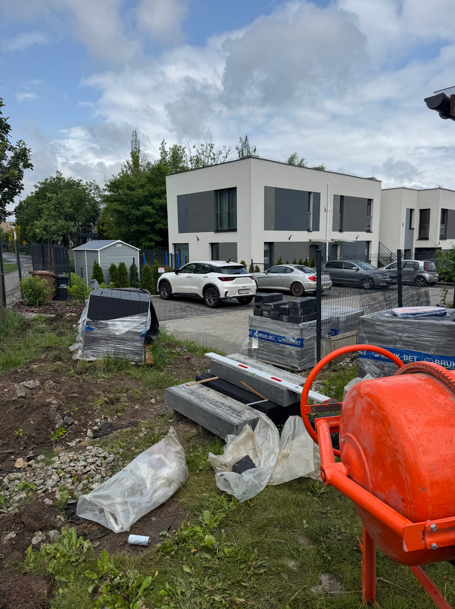 Przygotowanie do układania kostki brukowej: palety z kostką, obrzeża i betoniarka na tle nowoczesnych domów i aut. Widoczne ślady prac ziemnych.