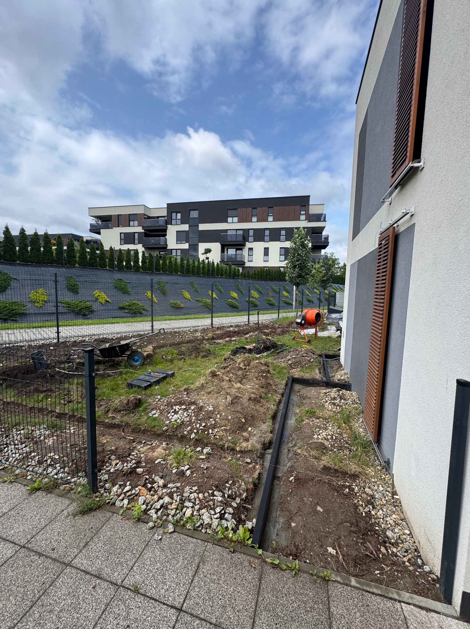 Przygotowanie terenu pod kostkę brukową: wykop, obrzeża, gruz. W tle budynek mieszkalny i ogrodzenie z roślinami. Widoczna betoniarka.