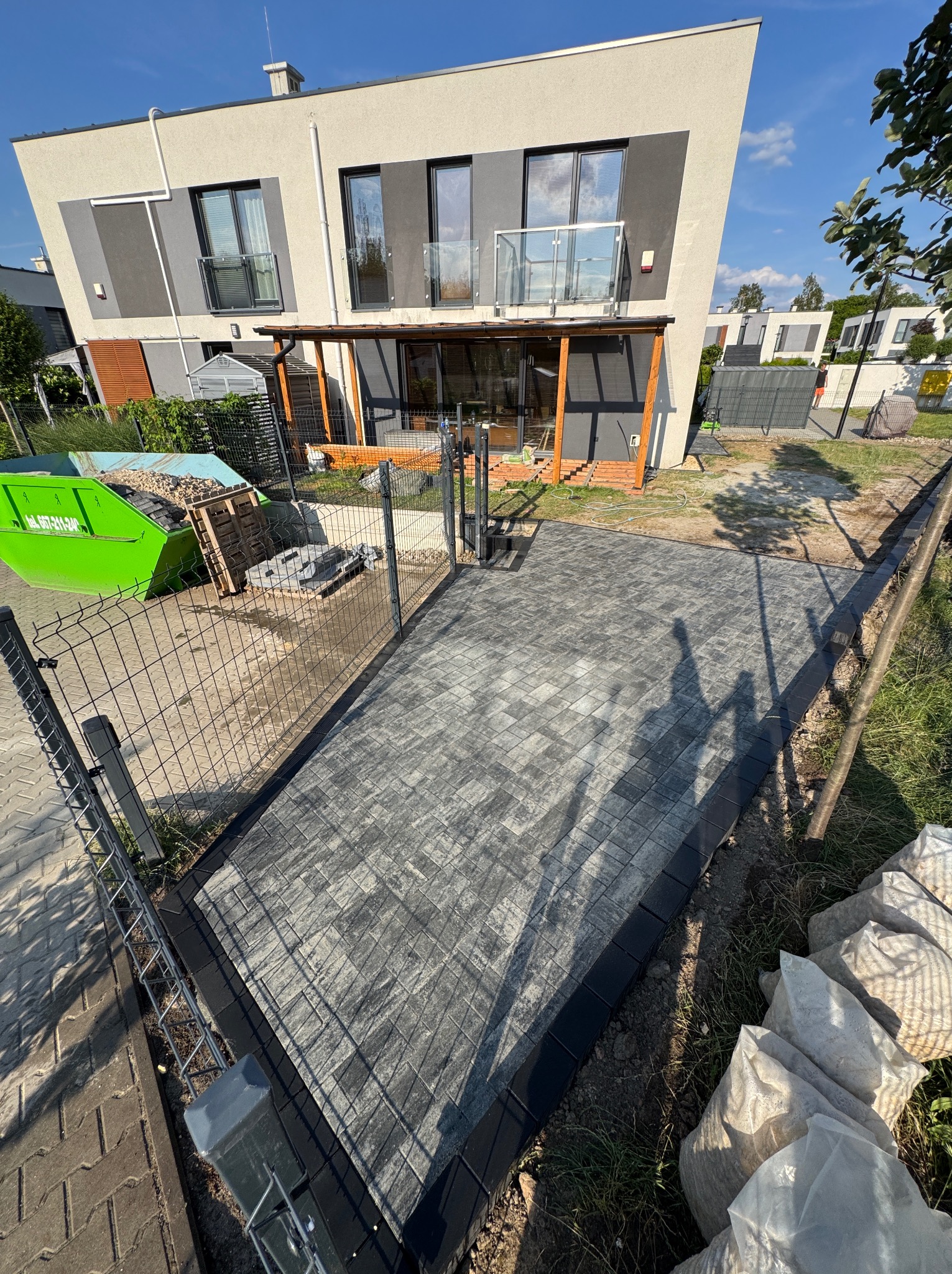 Nowo ułożona, szara kostka brukowa prowadząca do domu z drewnianym zadaszeniem, widoczny zielony kontener i worki z materiałem budowlanym.