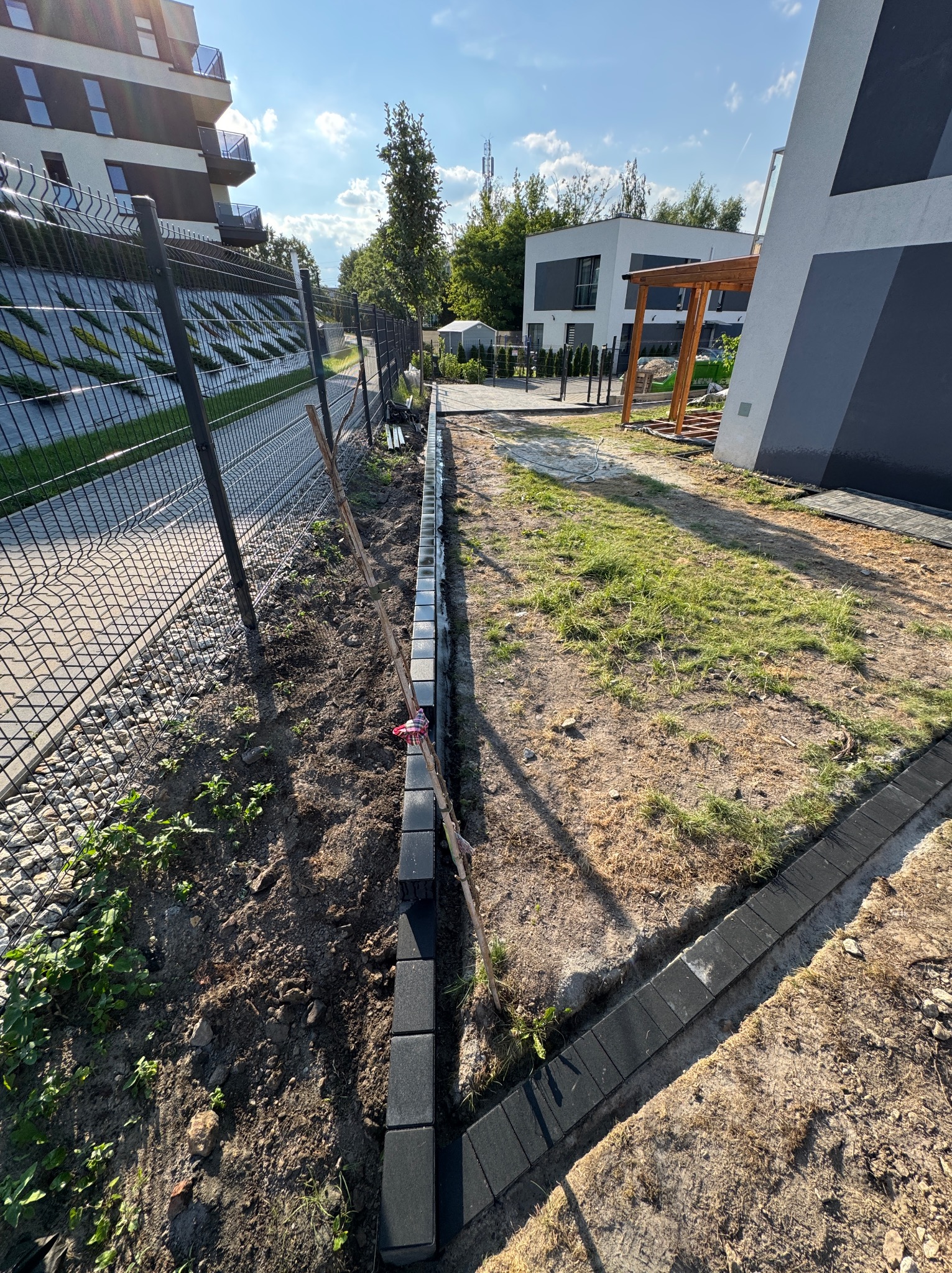 Fragment ogrodu w trakcie prac: obrzeża z kostki ułożone wzdłuż ogrodzenia, widoczne przygotowanie terenu pod dalsze prace. Nowoczesna zabudowa w tle.