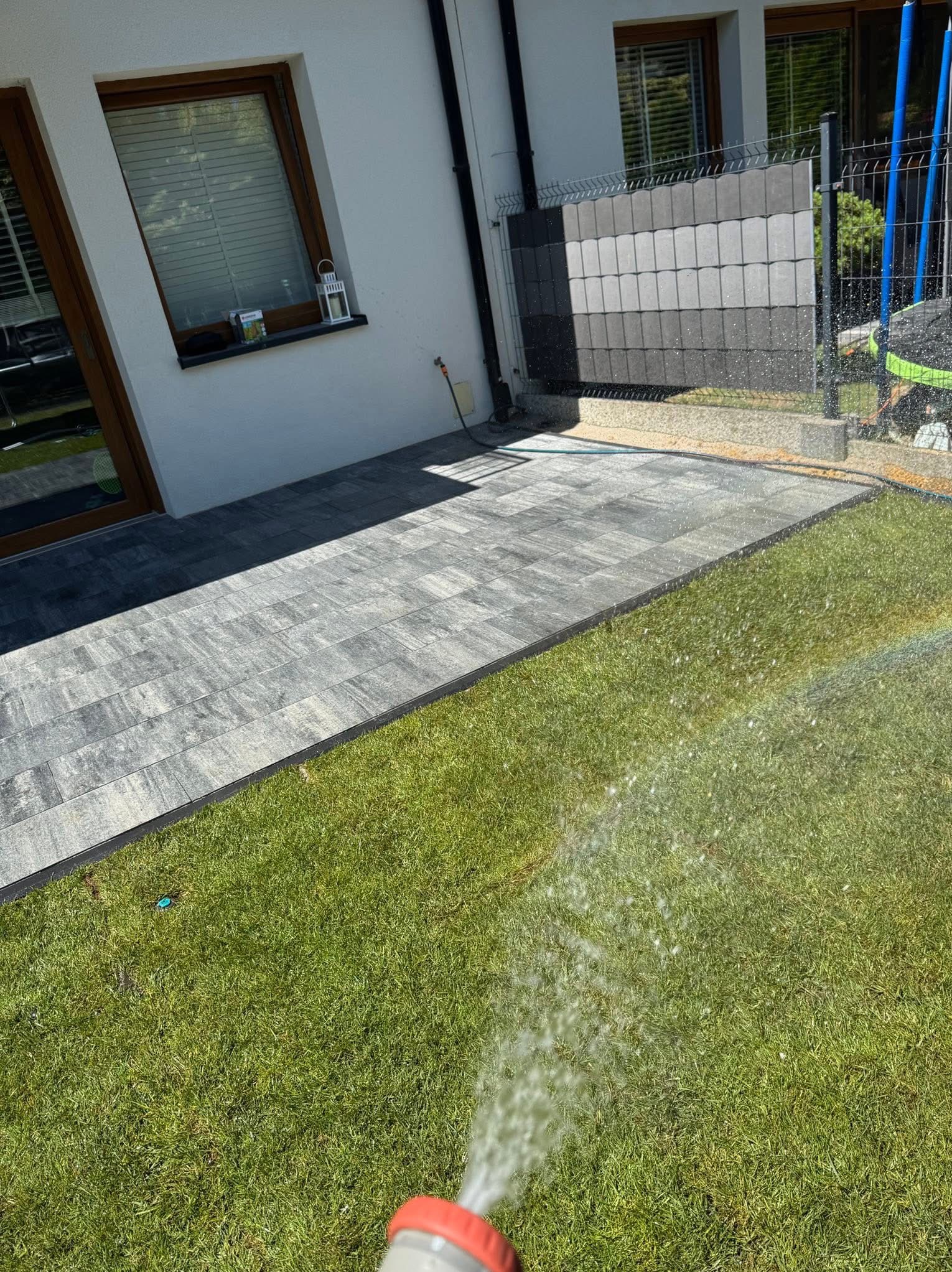 Szara kostka brukowa ułożona przy białym domu, obok trawnik zraszany wodą. Widoczny fragment ogrodzenia panelowego.