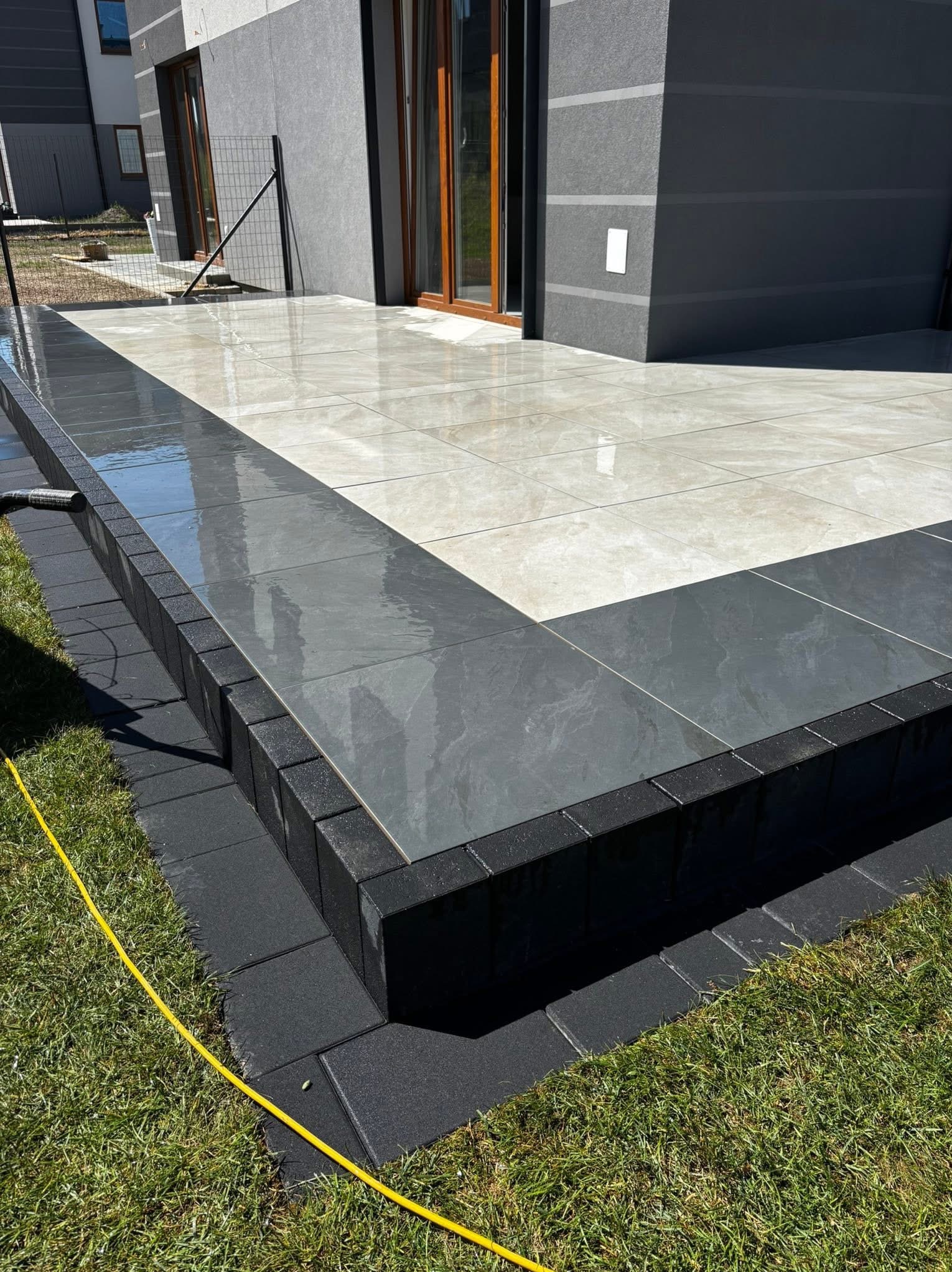 Nowoczesny taras z jasnych i ciemnych płytek, obrzeża z ciemnej kostki brukowej, widok na budynek z szarą elewacją i trawnik. Estetyczne wykończenie.