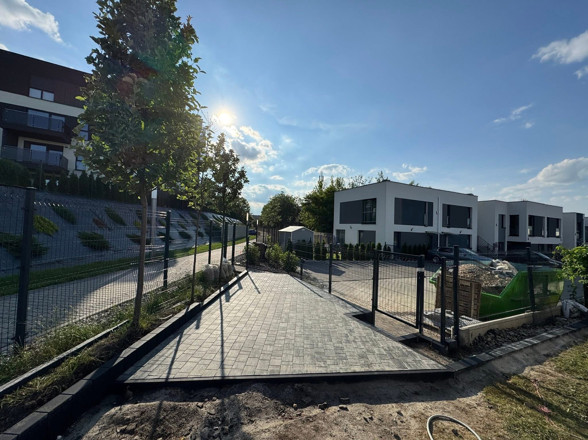 Nowa, szara kostka brukowa na podjeździe w Mikołowie, obramowana czarnym obrzeżem. Widok na nowoczesne domy i ogrodzenie panelowe w słoneczny dzień.