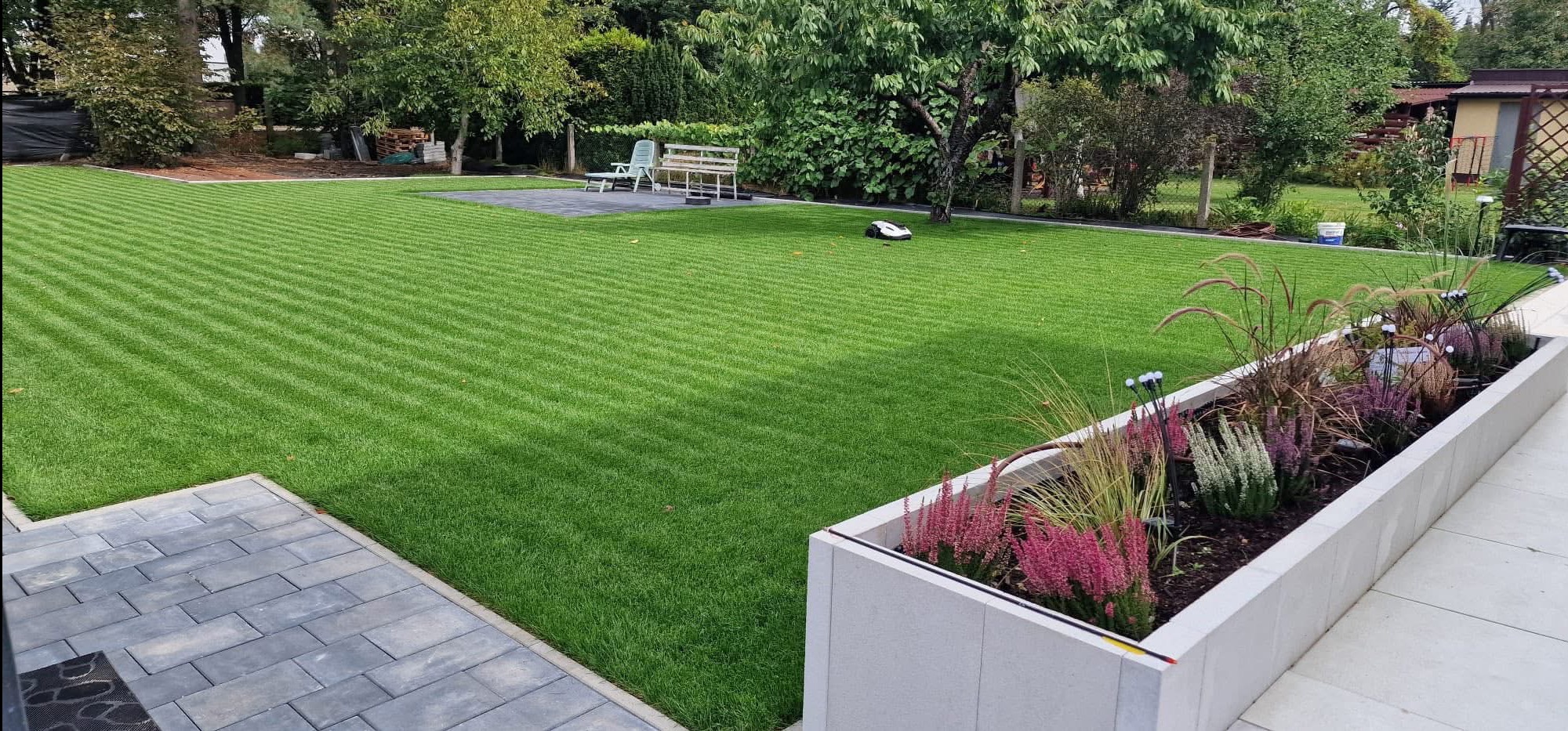 Perfekcyjnie skoszony trawnik z wyraźnymi pasami, rabata kwiatowa w betonowej donicy, robot koszący i kostka brukowa. Zadbany ogród z elementami małej architektury.