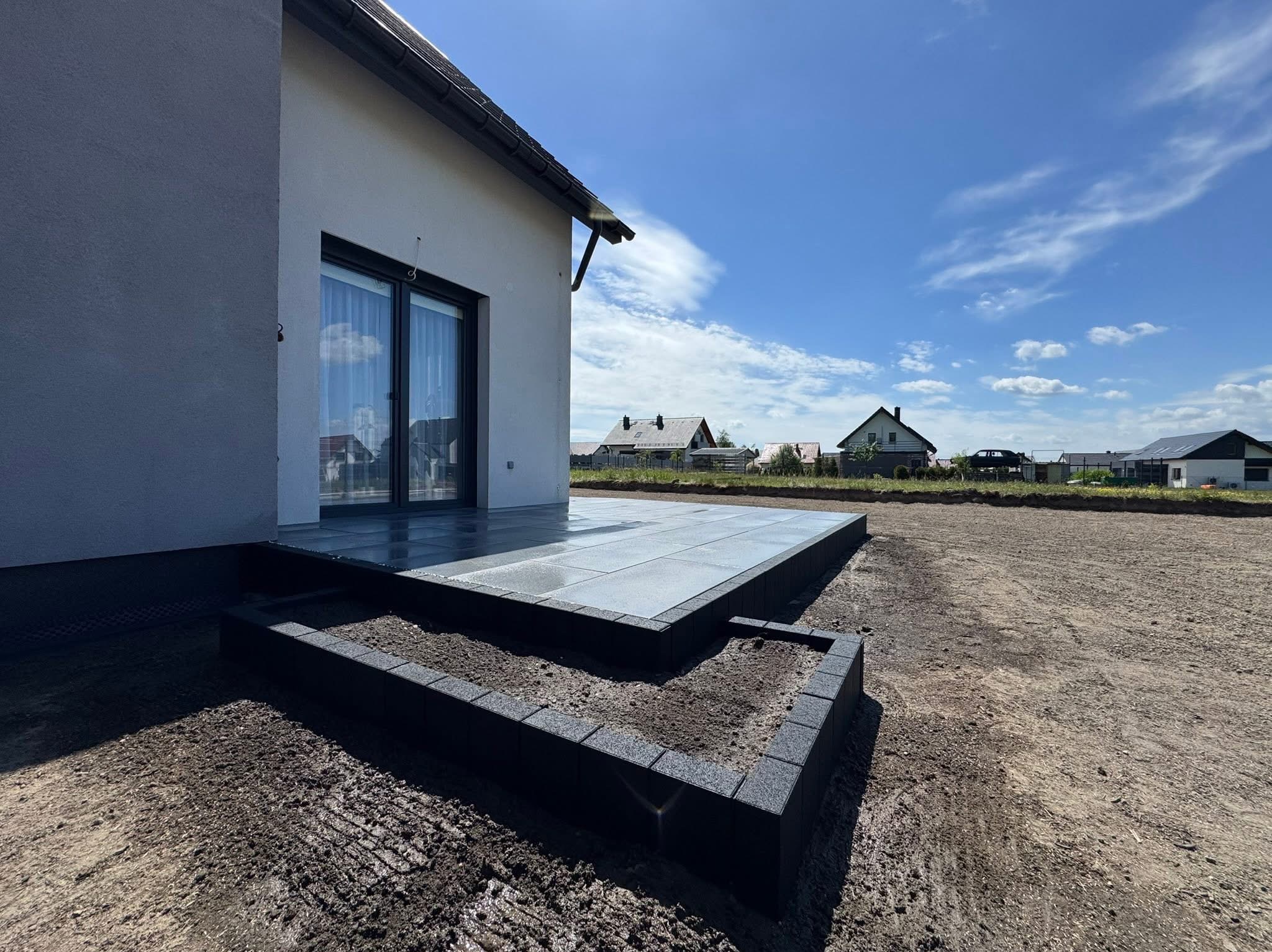 Nowoczesny taras z ciemnej kostki brukowej, schody z obrzeżami, przygotowany teren pod trawnik, dom jednorodzinny w tle, słoneczny dzień.