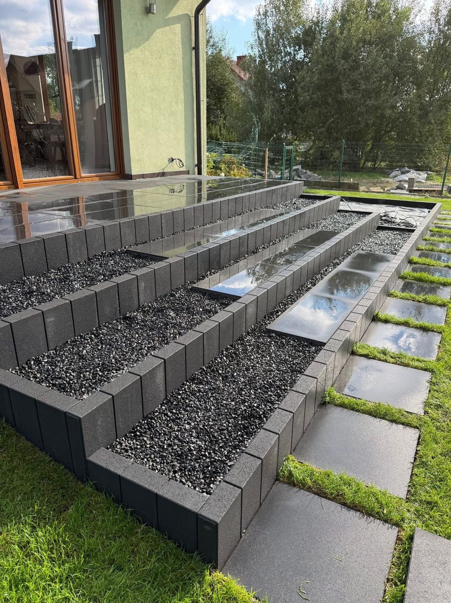 Nowoczesny taras z czarnych kostek i grysu, schodkowy układ. Elementy z trawy dodają naturalności. Widok na dom i ogród w tle. Po deszczu, mokra nawierzchnia.