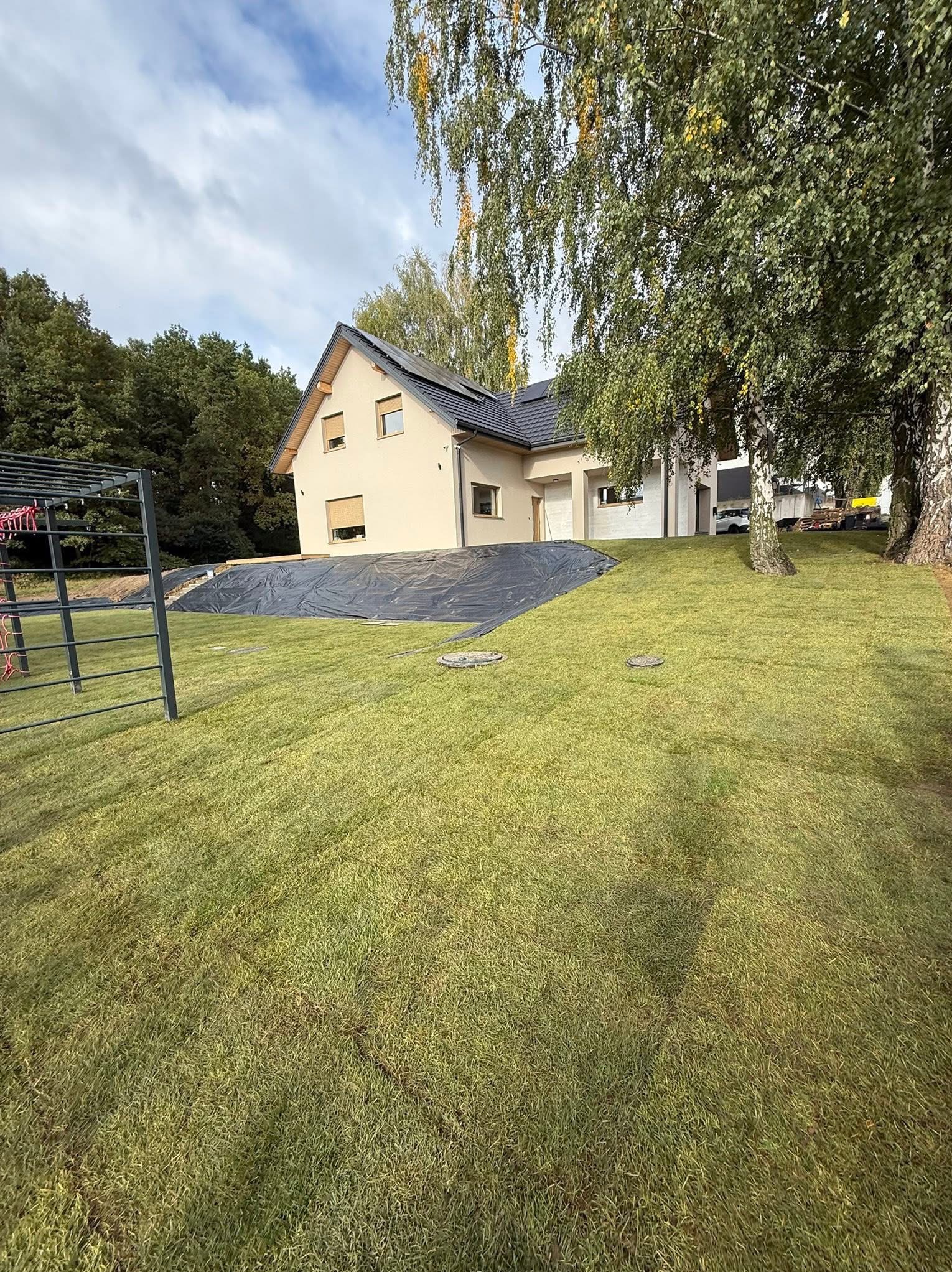 Nowo założony trawnik przed domem jednorodzinnym, z widocznymi drzewami i czarną folią zabezpieczającą skarpę. Świeża zieleń trawy kontrastuje z jasną elewacją budynku.