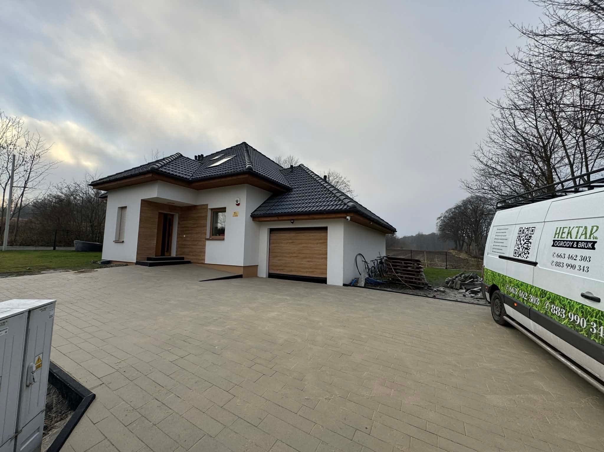 Nowoczesny dom z beżową kostką brukową na podjeździe. Na posesji stoi biały bus z logo firmy brukarskiej. Widoczne elementy wykończenia domu w kolorze drewna.
