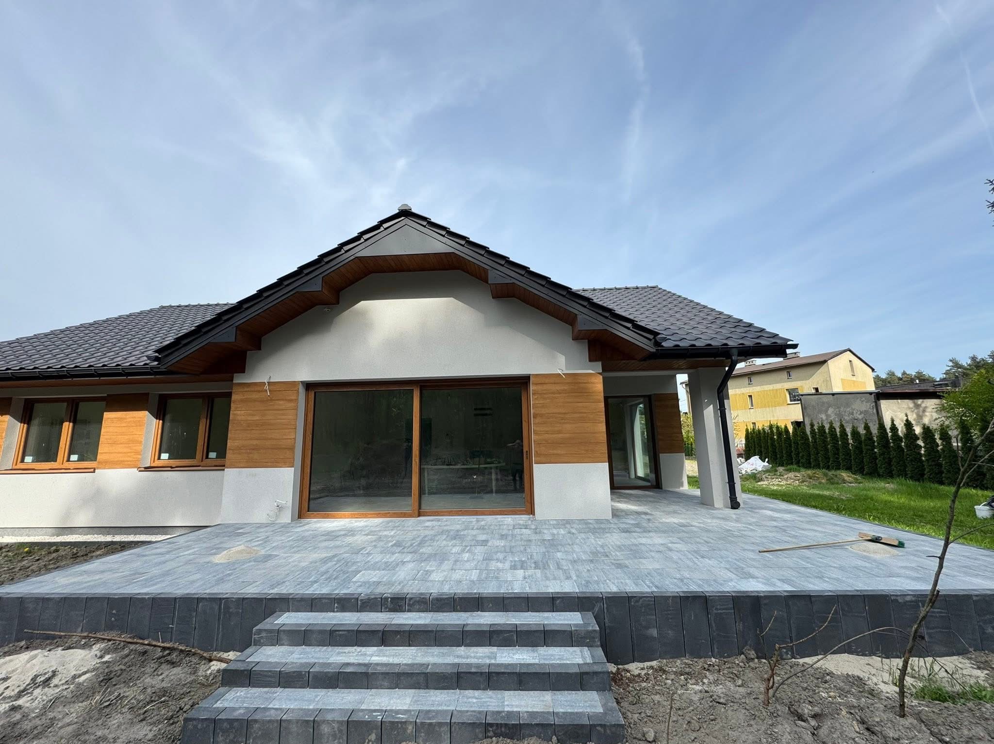 Nowoczesny dom z tarasem wyłożonym szarą kostką brukową. Widoczne schody z kostki, drewniane elementy elewacji i duże przesuwne okna tarasowe. Częściowo zagospodarowany teren.