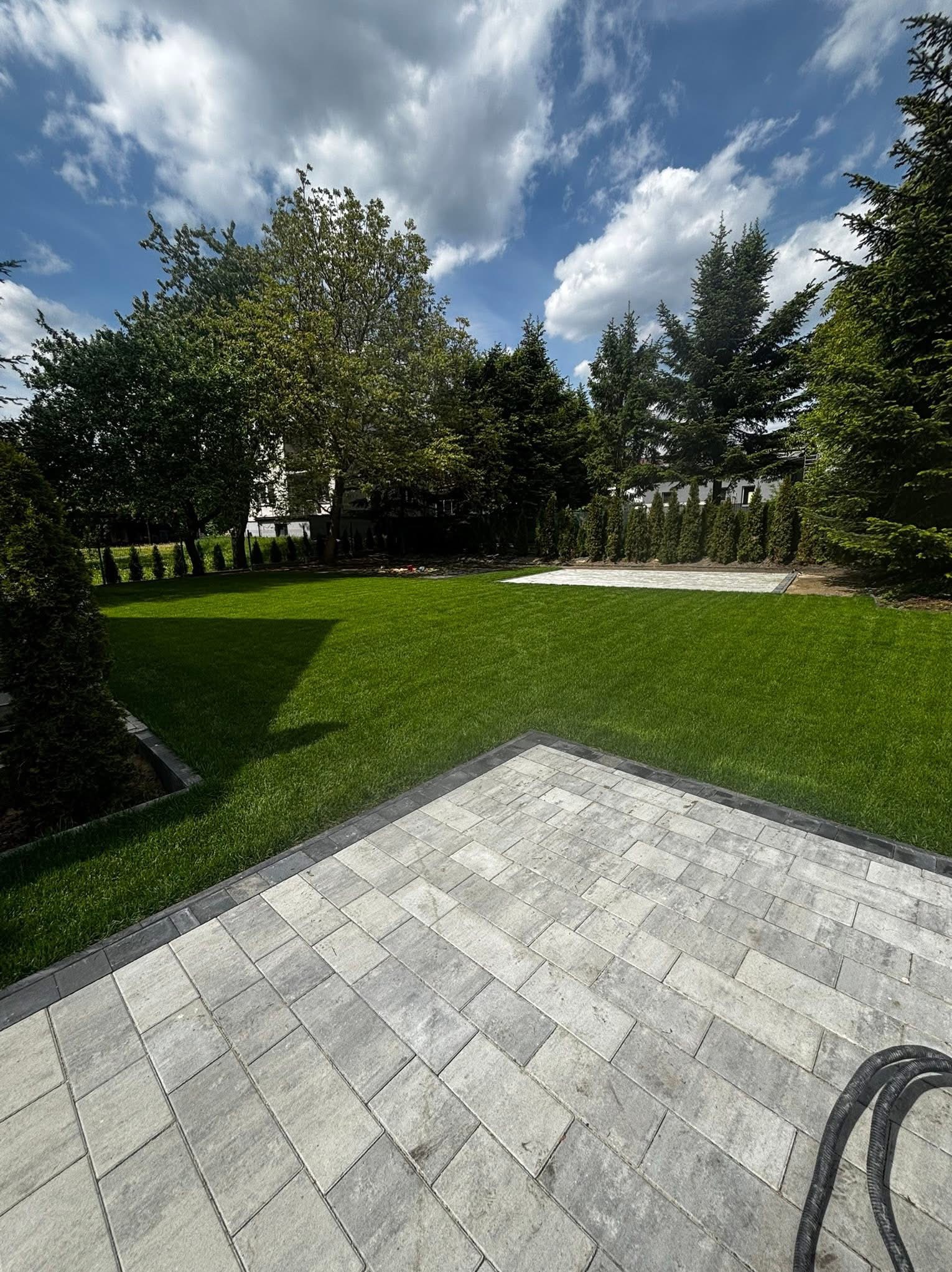 Ułożona kostka brukowa w odcieniach szarości, obrzeża z ciemnej kostki. W tle trawnik, drzewa iglaste i liściaste, błękitne niebo z chmurami. Słoneczny dzień.