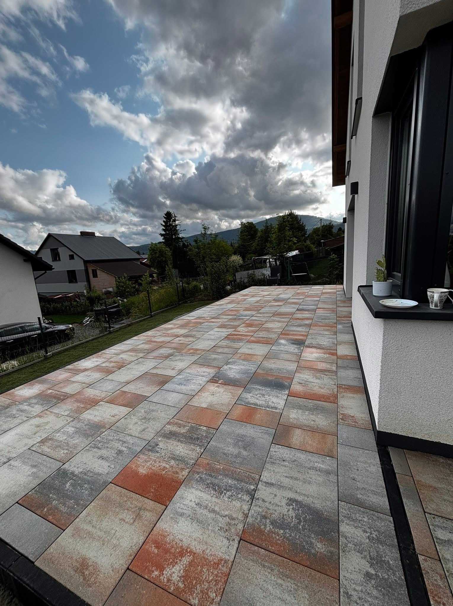 Nowoczesny taras z kostki brukowej w odcieniach szarości i czerwieni, przylegający do białego budynku z czarną stolarką okienną. W tle widok na góry i zachmurzone niebo.