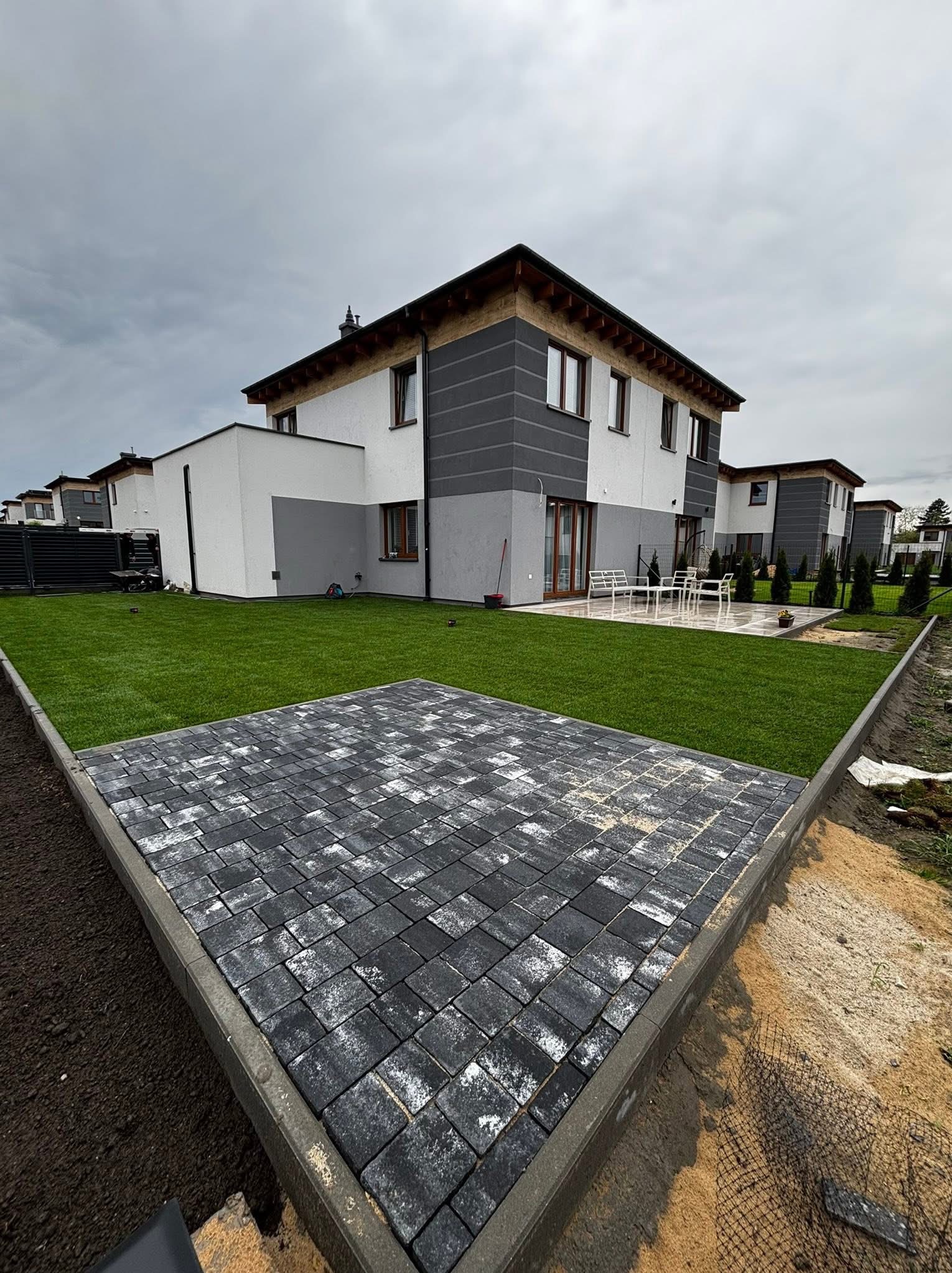 Nowoczesny ogród z równo ułożoną, szarą kostką brukową, obrzeżami i świeżą, zieloną trawą na tle domu w stonowanej kolorystyce. Pochmurne niebo.