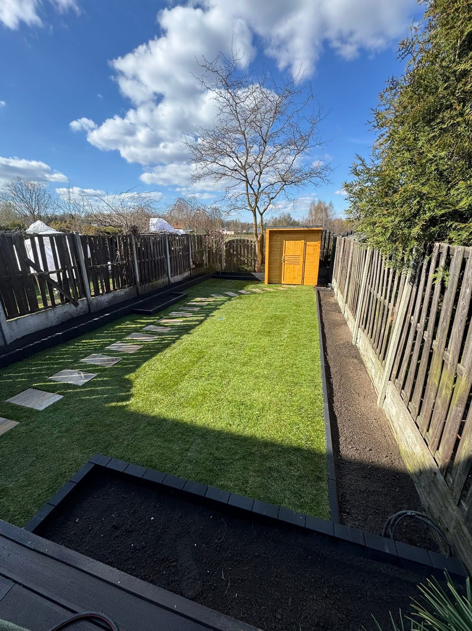 Zadbany ogród z równo przystrzyżoną trawą, ścieżką z płyt, drewnianym domkiem narzędziowym i rabatami wzdłuż ogrodzenia. Błękitne niebo z chmurami w tle.