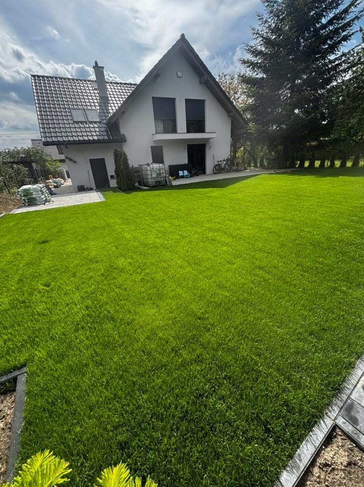 Świeżo założony trawnik z rolki przed nowoczesnym domem jednorodzinnym. Widoczne worki z ziemią i kostka brukowa na podjeździe. Intensywna zieleń trawy.