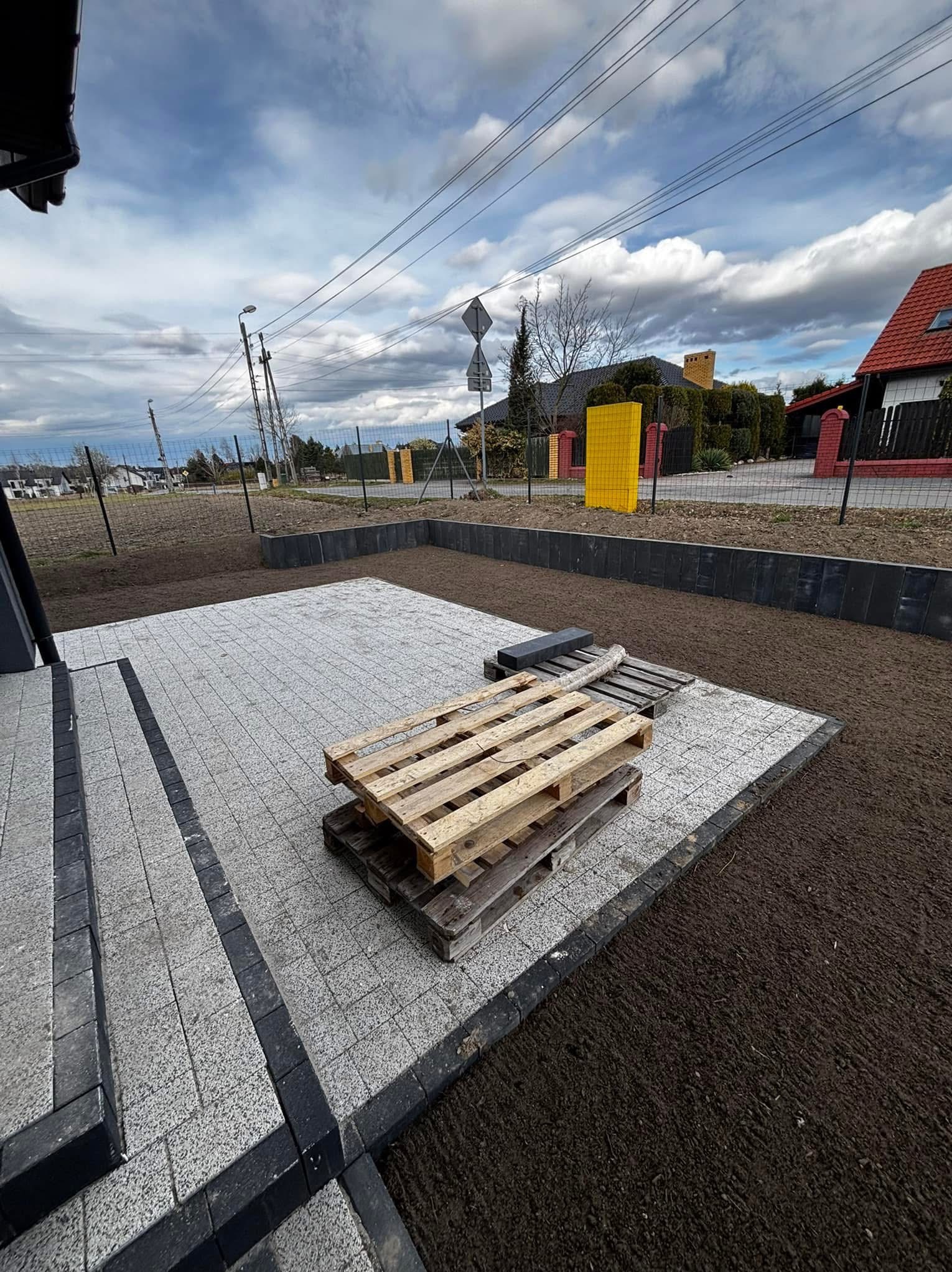 Ułożona kostka brukowa w szarym odcieniu z czarnym obramowaniem, palety drewniane na powierzchni, przygotowany teren pod trawnik, ogrodzenie w tle.