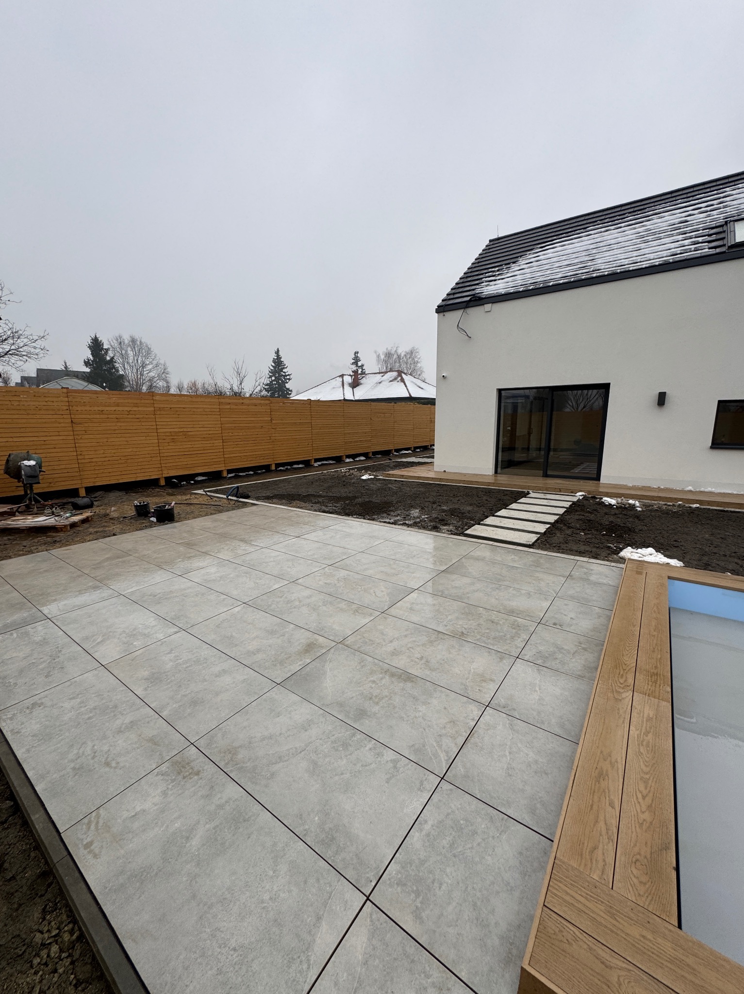 Nowoczesny taras z szarych płyt, drewniane obrzeże basenu, ścieżka z płyt prowadząca do domu. W tle drewniany płot i biały budynek w pochmurny dzień.