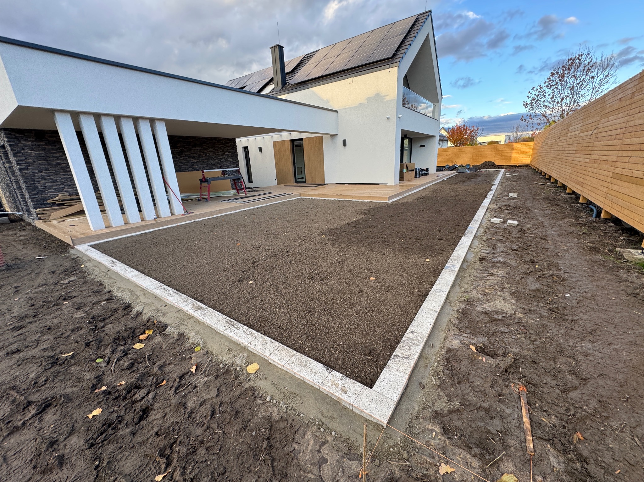 Przygotowany teren pod trawnik z obrzeżem z kostki przy nowoczesnym domu z panelami słonecznymi i drewnianym ogrodzeniem. Widoczne narzędzia i deski.