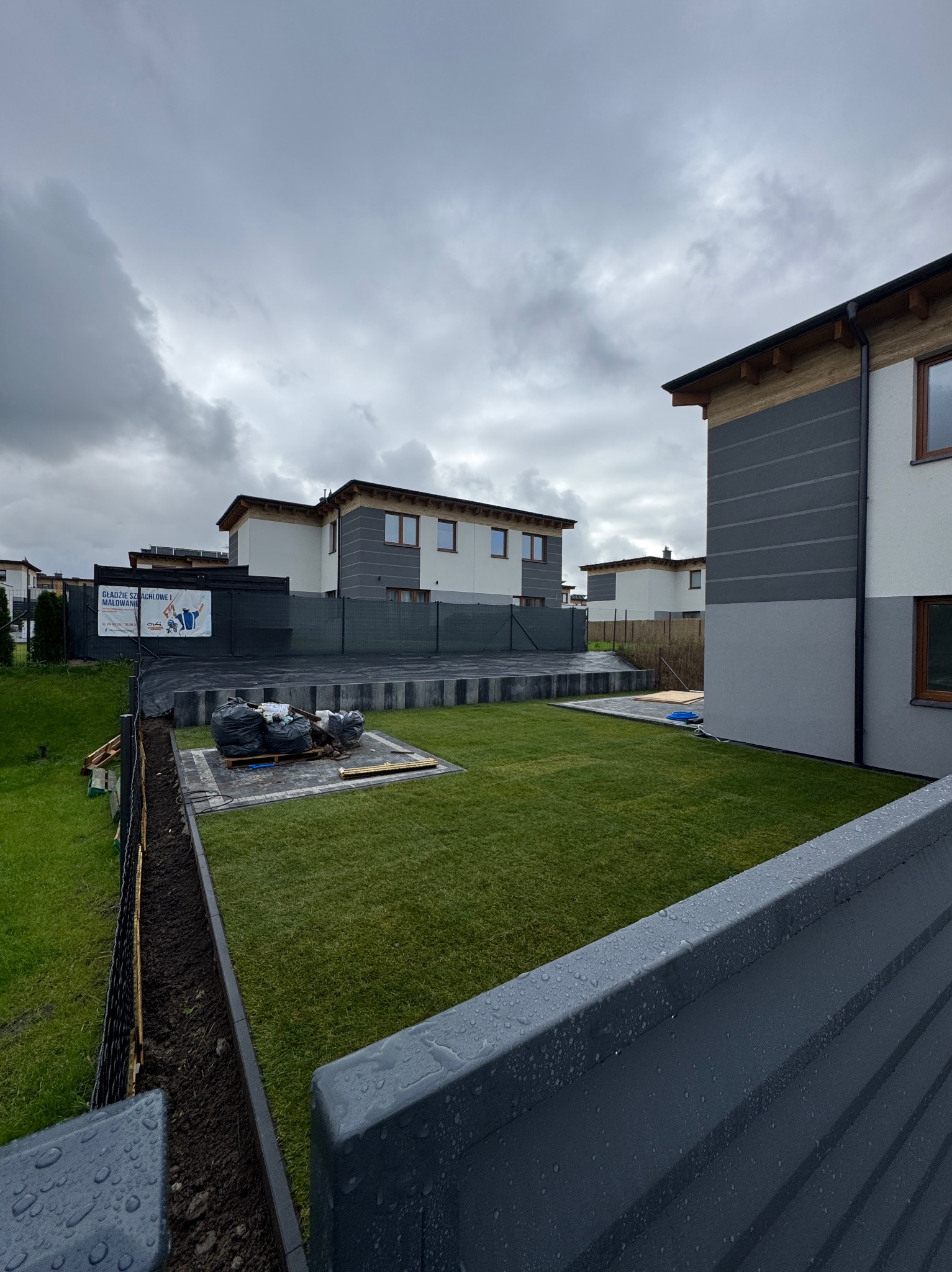 Świeżo założony trawnik z rolki na posesji z nowoczesnymi domami w zabudowie szeregowej, widoczne przygotowania do dalszych prac ogrodowych.
