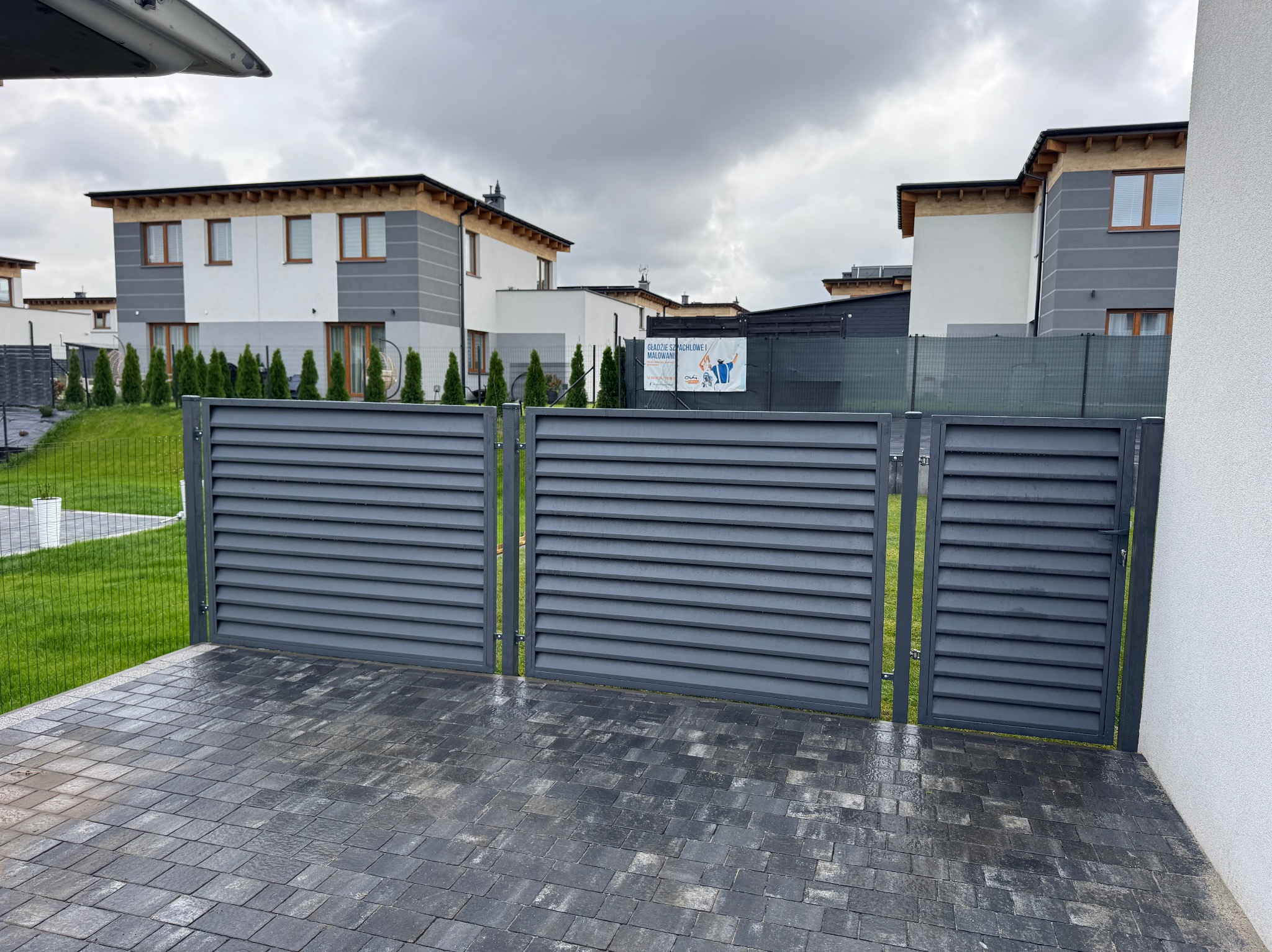 Nowoczesna, szara brama lamelowa z widokiem na posesję z kostką brukową i budynkami w tle, w pochmurny dzień. Minimalistyczny design i precyzyjne wykonanie.