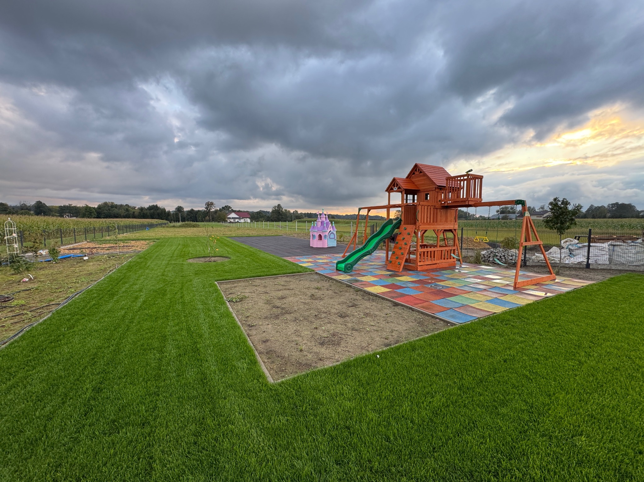 Nowo założony trawnik z rolki, plac zabaw z domkiem i zjeżdżalnią na kolorowej nawierzchni, w tle dom i pochmurne niebo. Widok z góry.