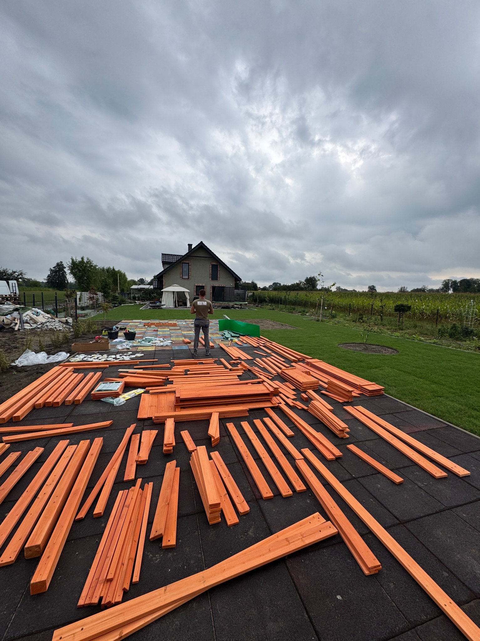Przygotowane elementy drewniane na czarnej, gumowej nawierzchni, prawdopodobnie do budowy konstrukcji ogrodowej. W tle dom i zielony trawnik pod pochmurnym niebem.