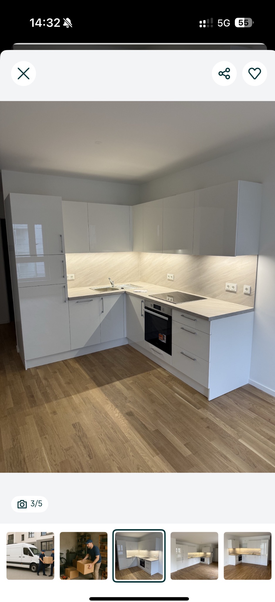 Biała, narożna kuchnia z połyskiem, oświetleniem podszafkowym i drewnianą podłogą. Minimalistyczny design, nowoczesne wykończenie. Widok na płytę indukcyjną i piekarnik.