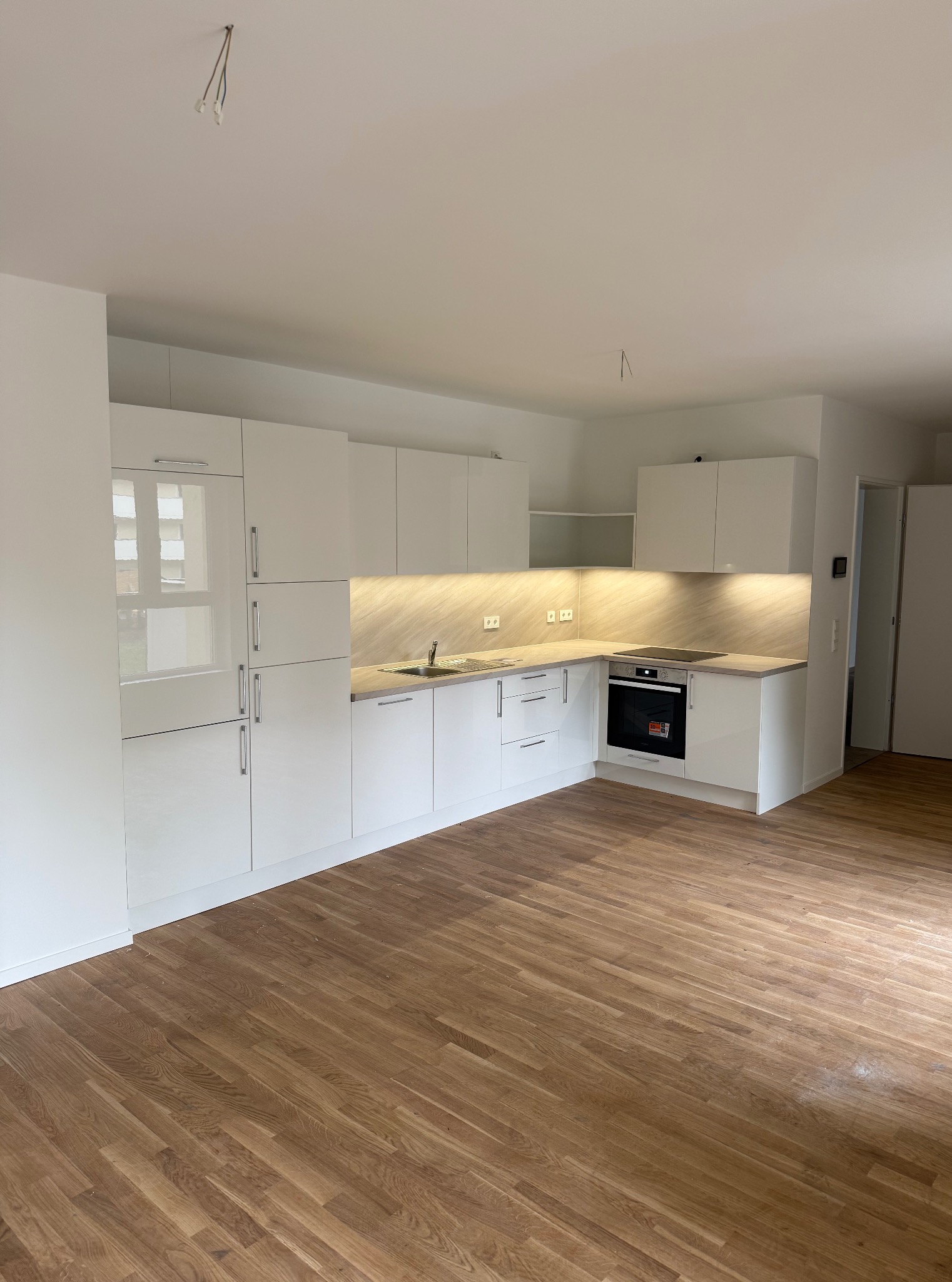 Biała, lśniąca kuchnia z drewnianą podłogą. Uwagę przykuwają minimalistyczne szafki i oświetlenie LED pod blatem, tworzące ciepłą atmosferę. Widoczne niedokończone podłączenie elektryczne.