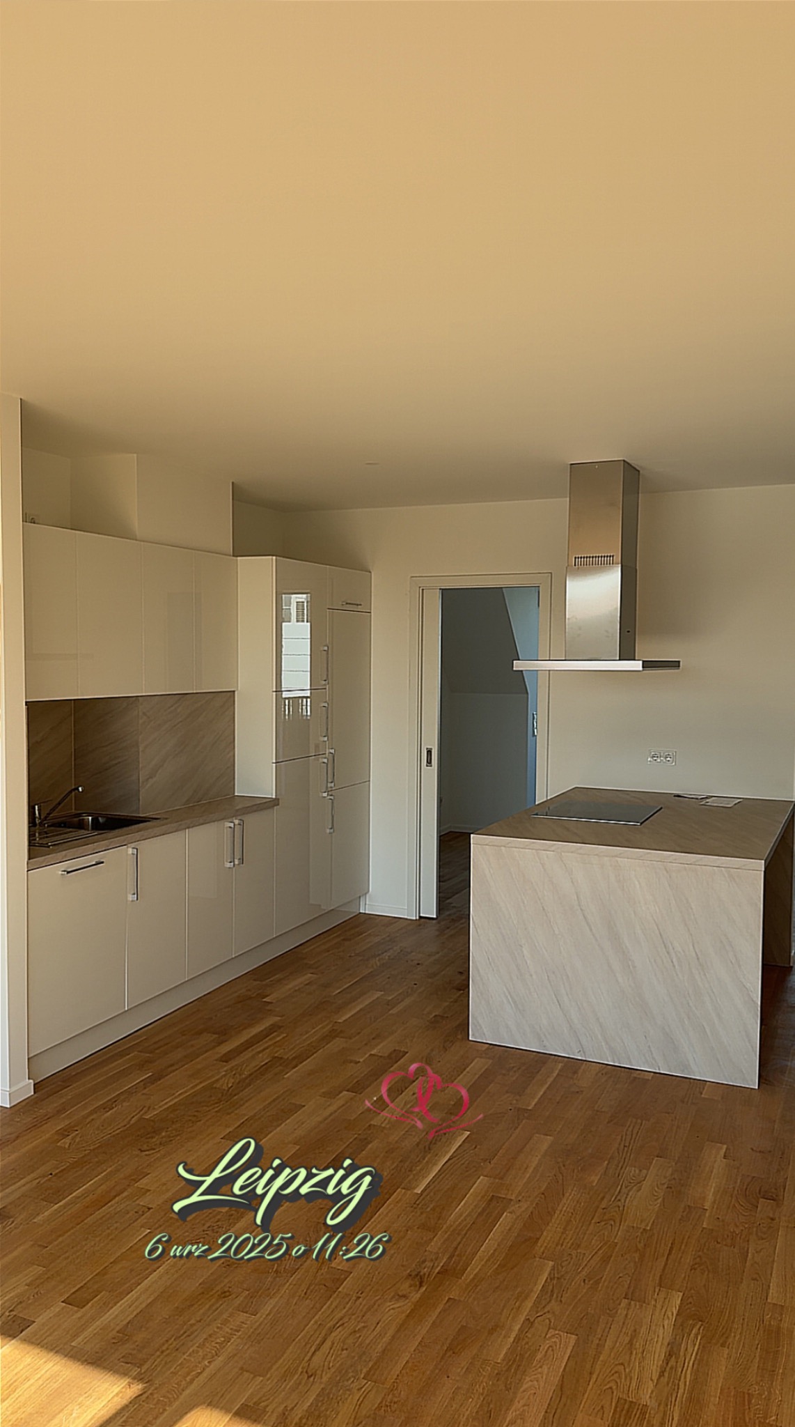 Jasna, nowoczesna kuchnia z wyspą i drewnianą podłogą. Białe, lakierowane fronty szafek i stalowy okap. Widoczne elementy zabudowy i płyta indukcyjna na wyspie.