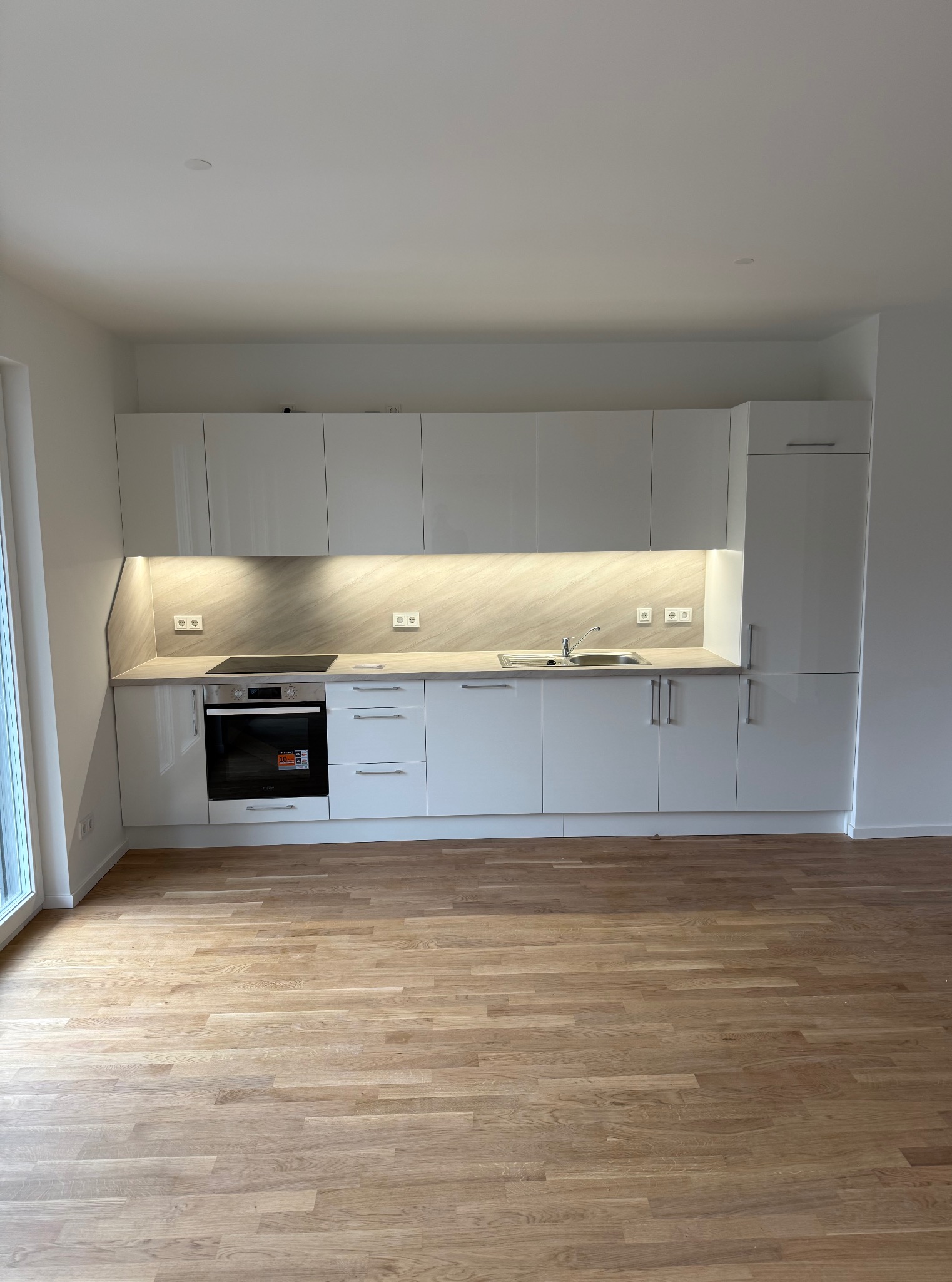 Biała, nowoczesna kuchnia z drewnianą podłogą. Błyszczące fronty szafek, oświetlenie podszafkowe, płyta indukcyjna i zlewozmywak. Minimalistyczny design, jasne wnętrze.