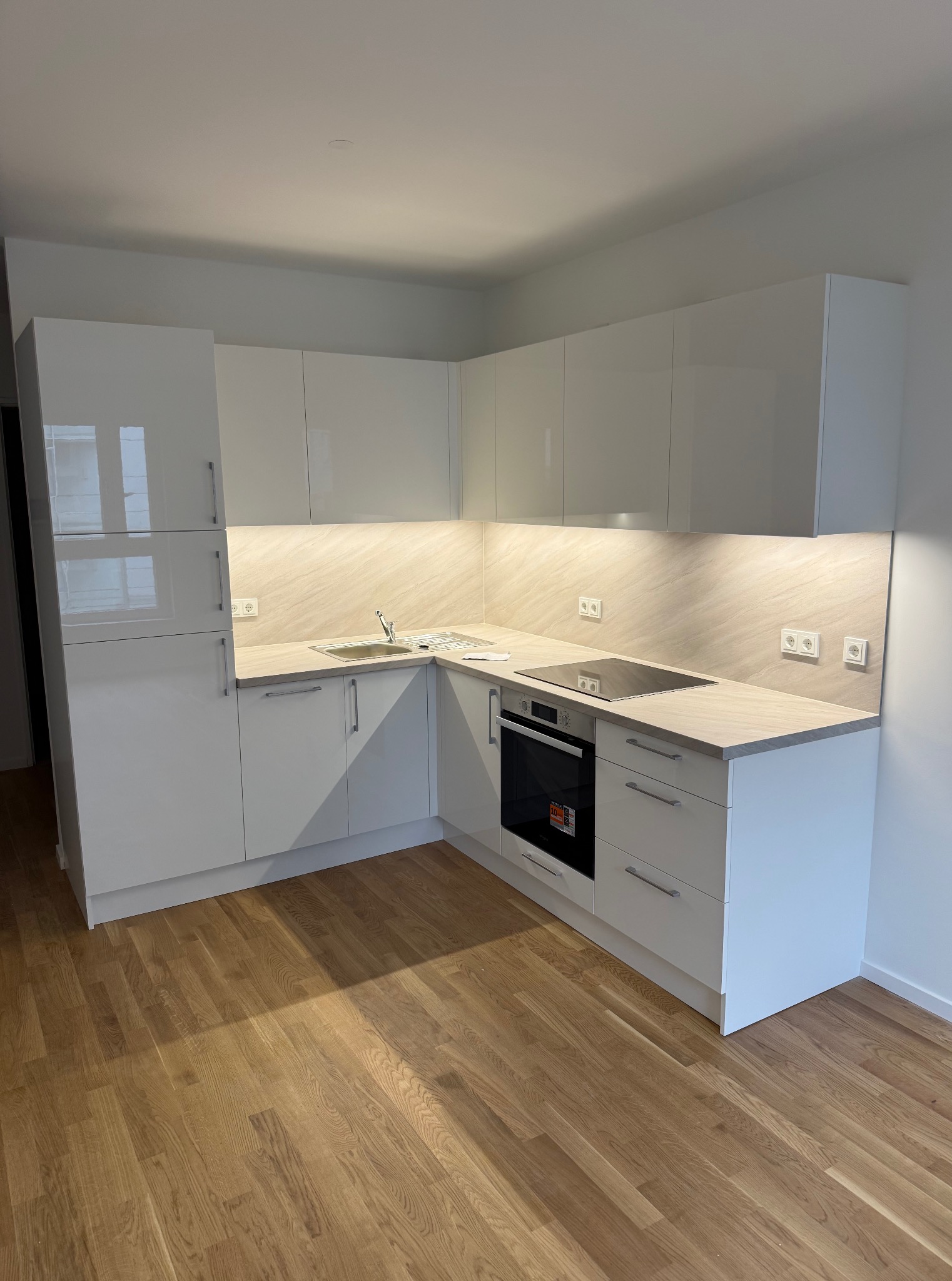 Biała, błyszcząca zabudowa kuchenna w kształcie litery L, z drewnianym blatem i podświetleniem LED, na tle parkietu. Widoczne gniazdka elektryczne.