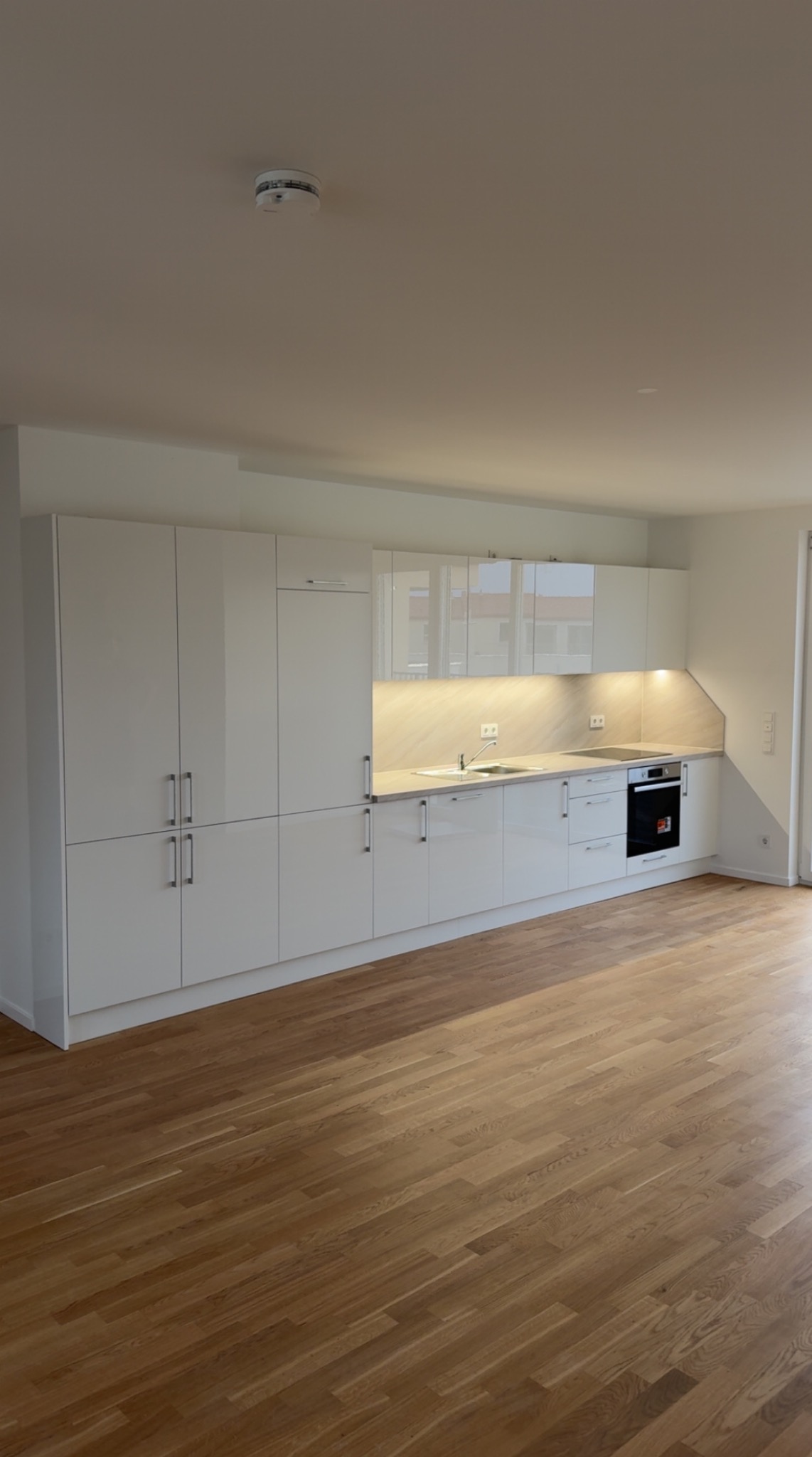 Nowoczesna, jasna kuchnia z białymi, lakierowanymi frontami szafek i drewnianą podłogą. Oświetlenie podszafkowe podkreśla blat. Widoczny czujnik dymu na suficie.